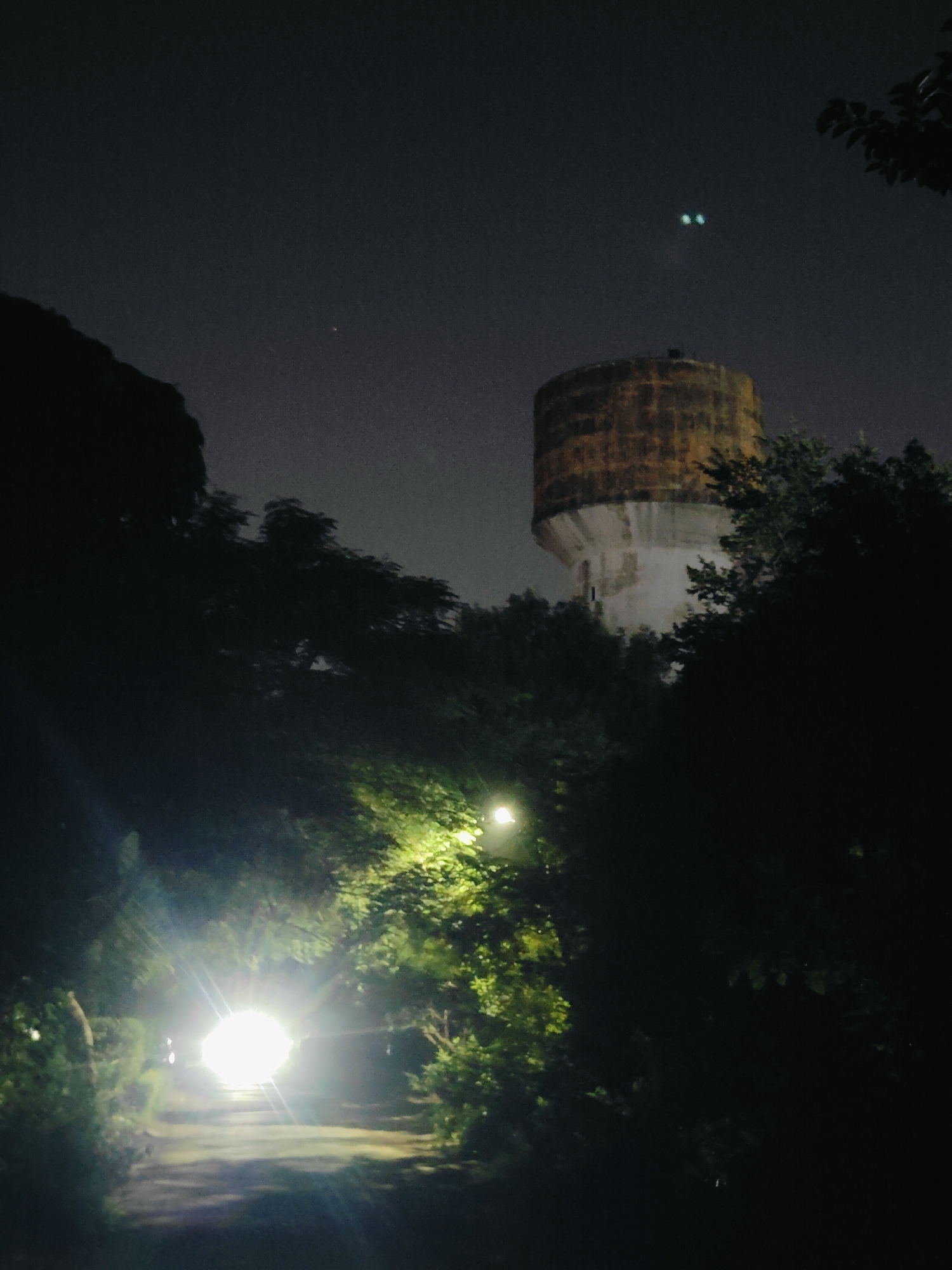 The image depicts a nighttime scene with a prominent water tower in the background, likely located in Gurgaon, India. The tower, a cylindrical structure with a rusted top and weathered concrete base, stands against a dark, cloudy sky. Two faint, greenish lights are visible in the sky above the water tower, possibly distant lights or celestial objects. In the foreground, a path is illuminated by a bright, intense light source, creating a strong glare and washing out the surrounding foliage. Dense trees and vegetation are visible on either side of the path, their leaves silhouetted against the dim light. The overall atmosphere is dark and somewhat mysterious, with the artificial light contrasting sharply with the natural darkness. There are no visible people or animals in the image.