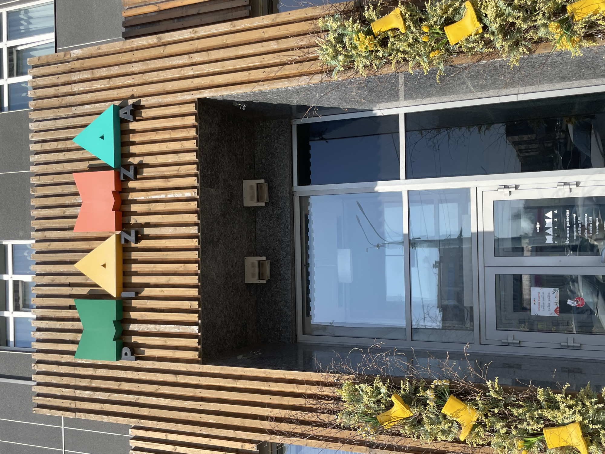 The image shows the facade of a building, possibly a cafe called "Mama," located in Izhevsk, Russia. A wooden slat structure forms the exterior wall, adorned with colorful geometric shapes spelling out "PIZZA." The entrance features glass doors and windows, reflecting the surrounding buildings. Planters filled with greenery and yellow rubber boots decorate the exterior. Posters with opening hours are visible on the door.