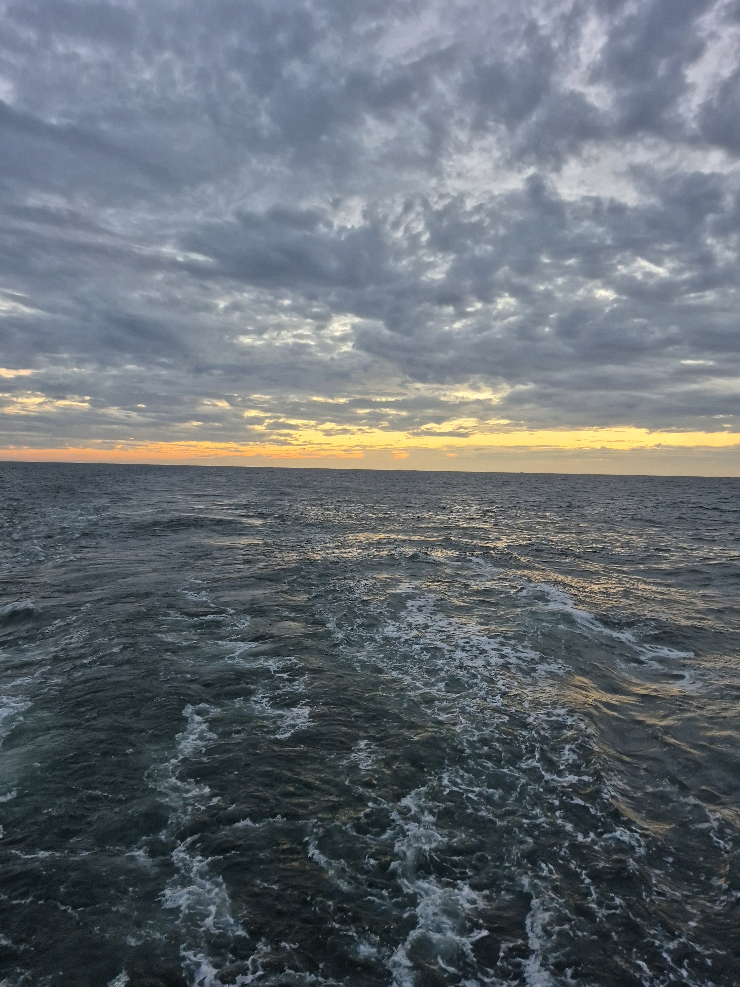 The image captures a wide-angle view of the sea under a cloudy sky. The horizon line is clearly defined, separating the choppy, dark blue water from the overcast sky. A band of warm orange and yellow light is visible just above the horizon, suggesting either sunrise or sunset. The clouds are a mix of gray and white, with some areas appearing darker and more dense than others. The water's surface is disturbed, with white foam and ripples indicating movement, likely from a boat or wind. The scene is devoid of any man-made structures or identifiable landmarks, making it impossible to determine a specific location within Unknown, Germany. There are no people or animals visible in the frame.