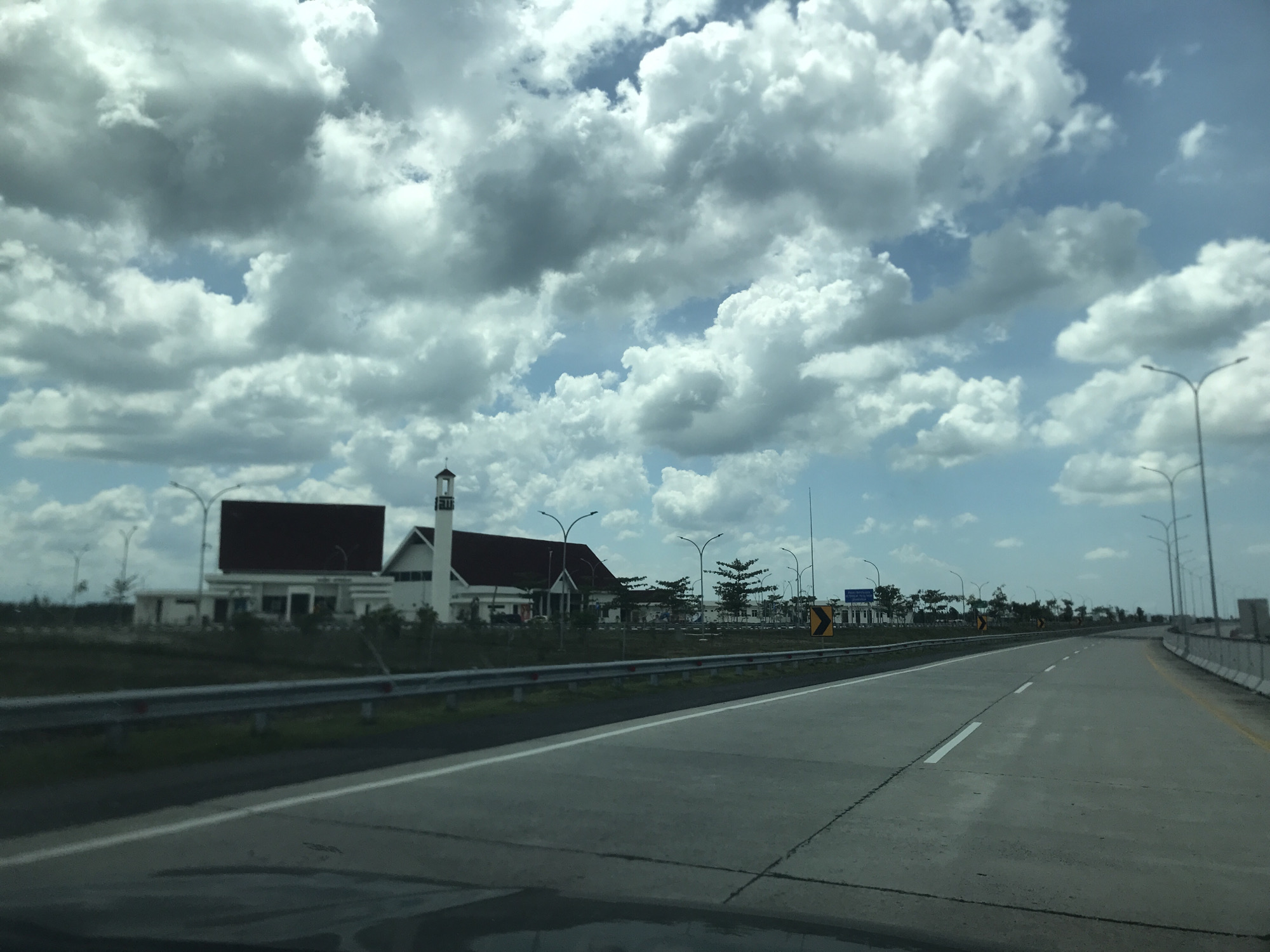 The image shows a view from inside a vehicle driving on a multi-lane highway in Gohor Lama, Indonesia. The road is made of grey concrete. The sky is filled with white cumulus clouds with some patches of light blue sky visible. To the left of the road, there is a long, white building complex with red rooftops and a tower that resembles a minaret. There is a metal guardrail along the edge of the road. Street lamps line the side of the road. There are other vehicles on the road.