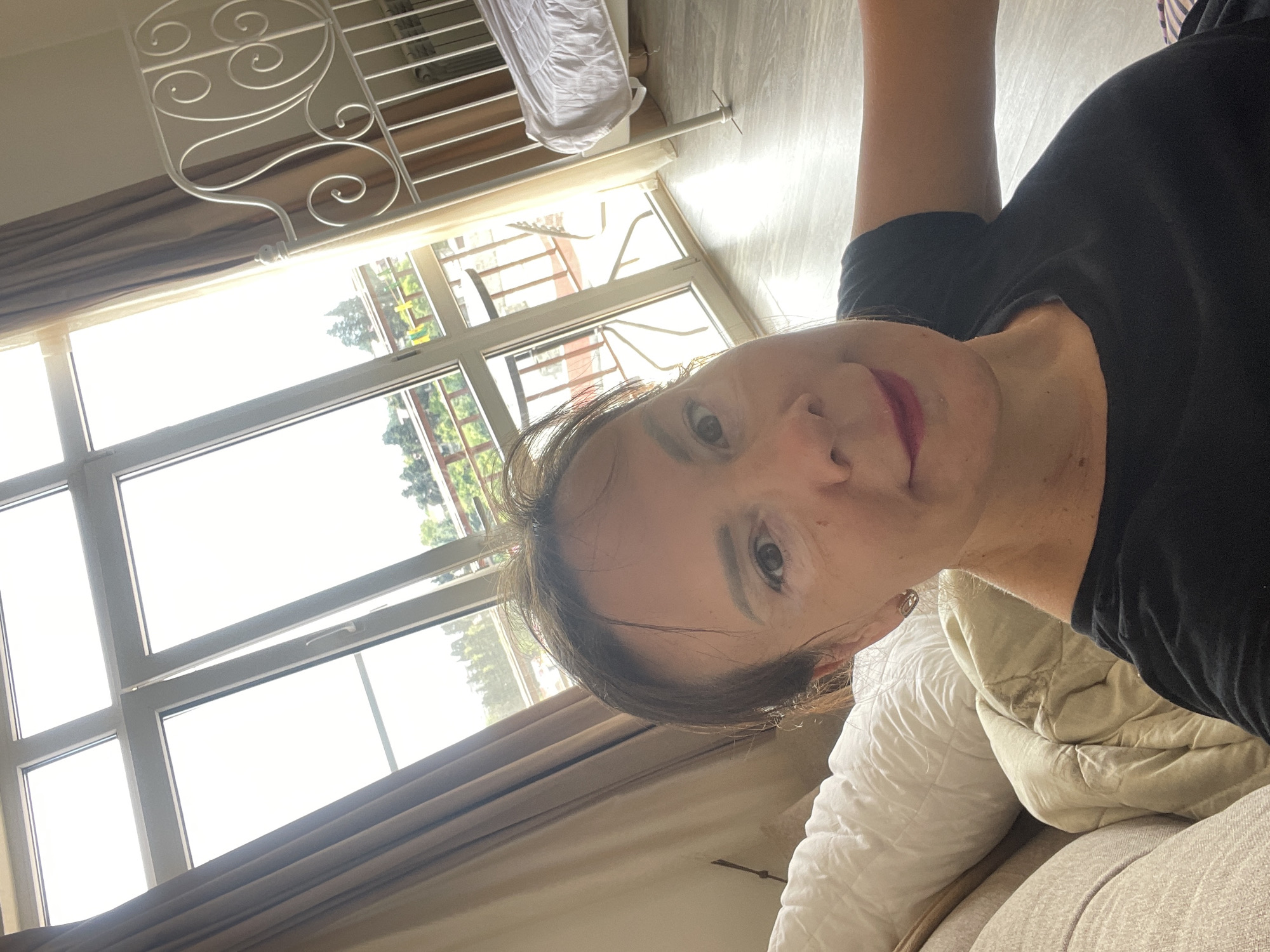 A selfie of a woman with shoulder-length brown hair and makeup is taken in what appears to be a bedroom in Sochi, Russia. She is wearing a black t-shirt and is seated on a sofa with pillows and blankets. Behind her, a large window with a balcony is visible, with curtains on the left. Outside the window, trees and buildings can be seen. To the right, a white metal bed frame with a white mattress and bedding is present. The room has light-colored wooden flooring.