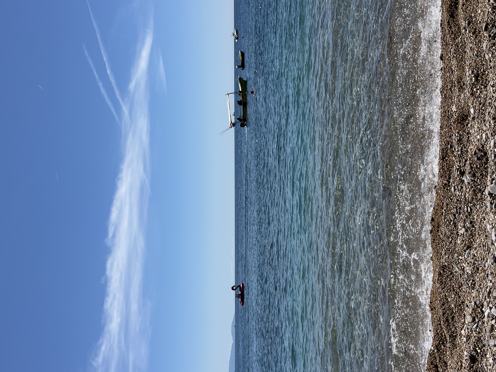 The image captures a coastal scene near Budva, Montenegro, showing a beach with small pebbles on the right. Clear water extends from the beach into the sea, with waves visible on the shore. In the distance, several boats are anchored in the water, including one that appears to be a fishing boat. A red inflatable boat with a person is visible in the water. The sky is blue with scattered white contrails from planes. The angle suggests the photo was taken from a position on the beach.