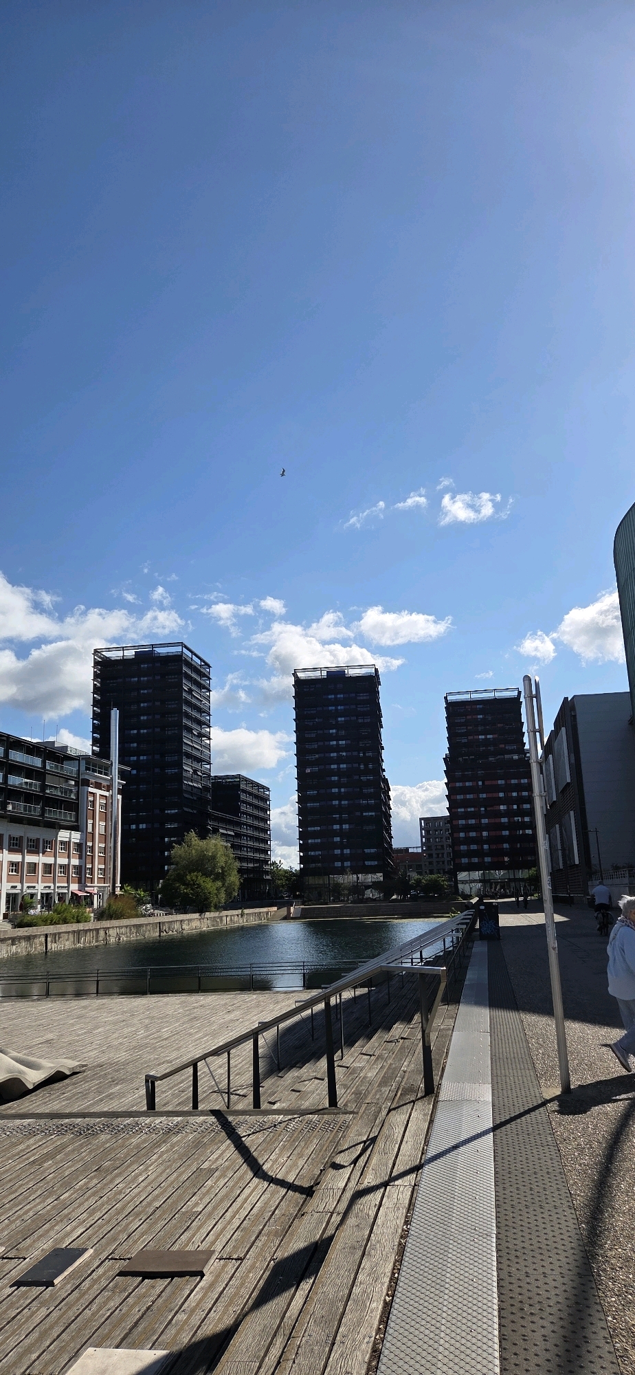 The image captures a waterfront view of modern high-rise buildings in Strasbourg, France, specifically in the area of the European Quarter. In the foreground, a wooden promenade with metal railings leads down to a calm body of water. The water reflects the sky and the surrounding architecture. Several tall, dark-colored residential buildings dominate the midground. The sky is a clear blue with scattered white clouds, and a single bird is visible in flight high above. To the right, a paved walkway with tactile paving is present, and a person is walking away from the camera. The overall scene suggests an urban environment with a focus on contemporary architecture and waterfront development.