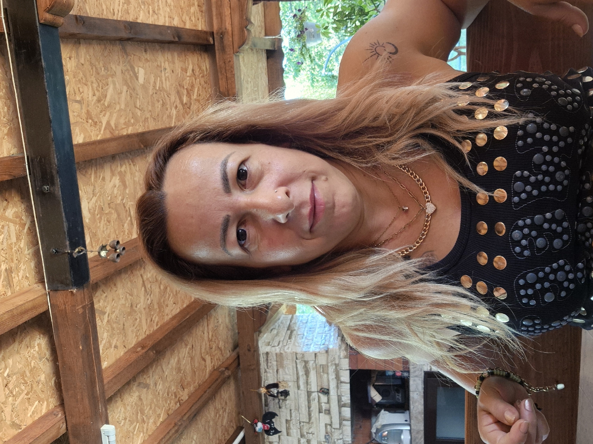 A medium shot portrait of a woman in her 40s with light brown hair, a nose ring, and light makeup. She is wearing a black tank top adorned with gold and grey circular studs. She has several gold necklaces, one with a heart-shaped pendant. A beaded bracelet is visible on her left wrist. A tattoo of a crescent moon and sun is visible on her right shoulder. The background is an outdoor covered patio with a wooden beam ceiling. To the left, there is a stone fireplace and kitchen appliances. To the right, lush greenery is visible. The woman is looking directly at the camera with a slight smile.