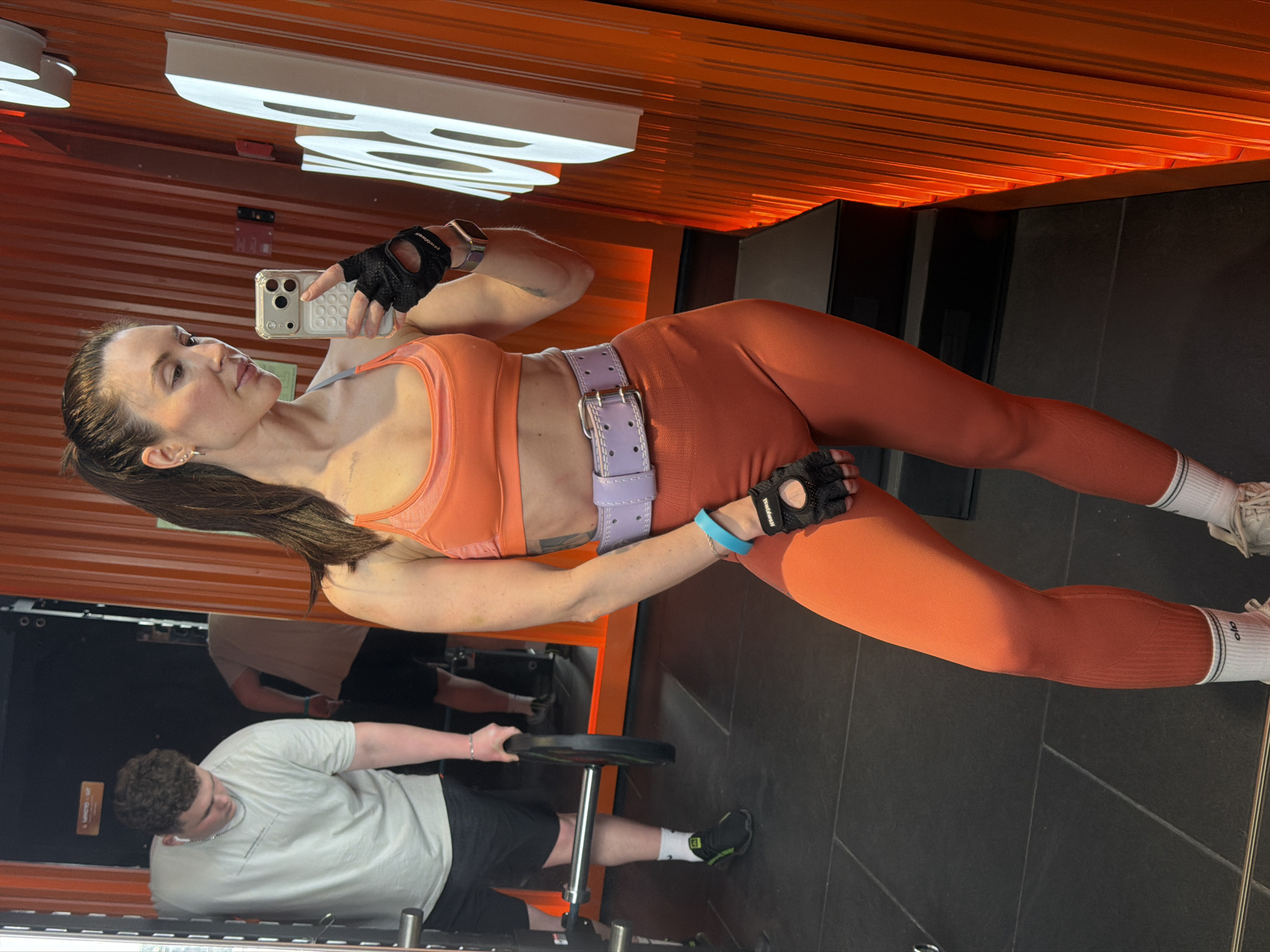 The image depicts a woman in workout attire taking a selfie in what appears to be a gym setting. She is wearing an orange sports bra, orange leggings, a purple belt, and workout gloves. She is holding a phone in her left hand, presumably to take the selfie. In the background, a man is visible near workout equipment, possibly lifting weights. The gym has an orange and black color scheme. While the city of Perm, Russia, cannot be verified, the image suggests an indoor fitness facility.