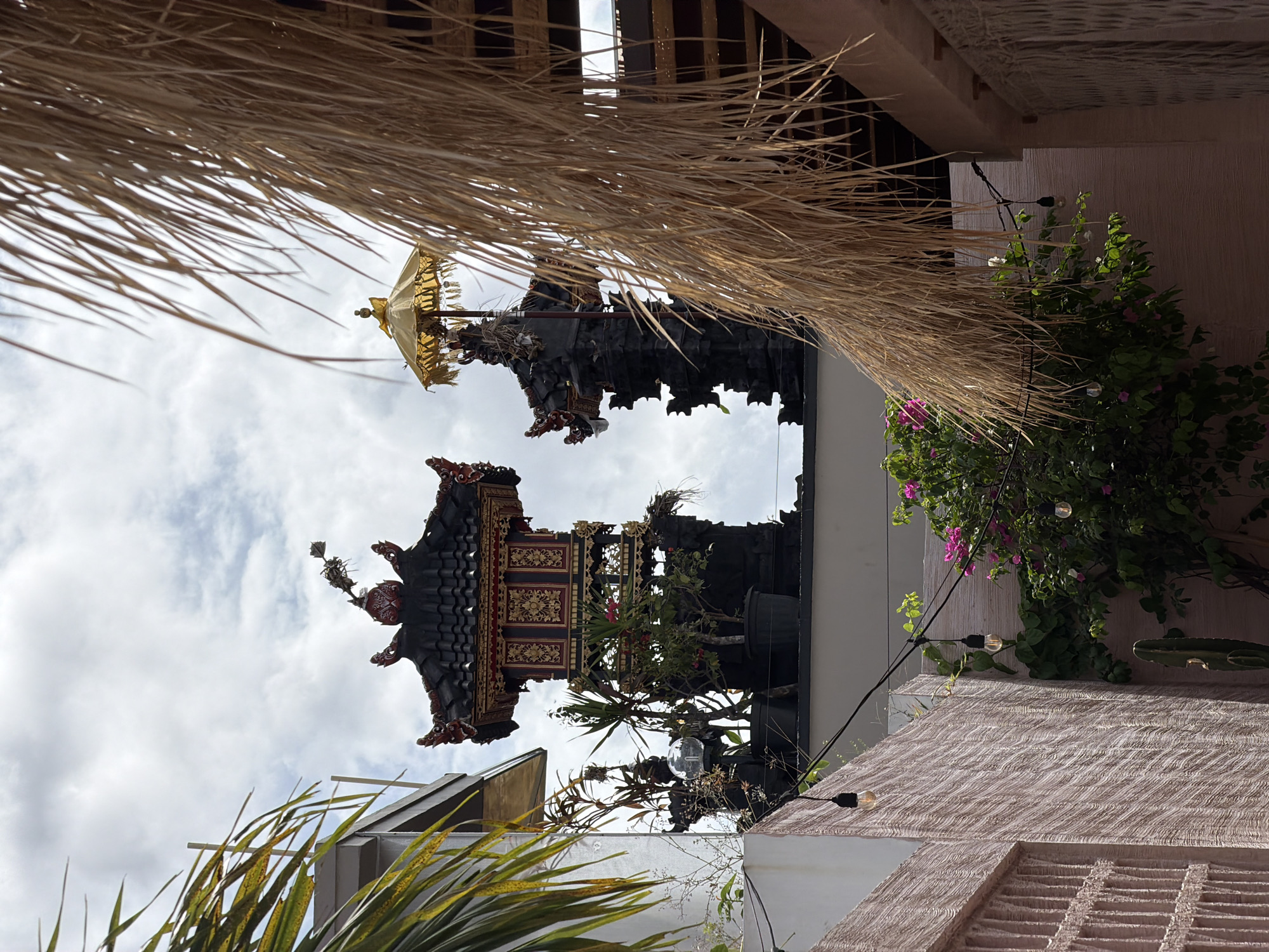 The image shows a traditional Balinese structure with a multi-tiered roof, likely a shrine or temple, viewed from a low angle, possibly in North Kuta, Indonesia. The structure has ornate carvings and decorations, including what appears to be a golden umbrella at the top. The sky is visible in the background, with some clouds. In the foreground, there is a thatched roof and some vegetation, including a plant with pink flowers. String lights are present against a textured wall with a cactus.