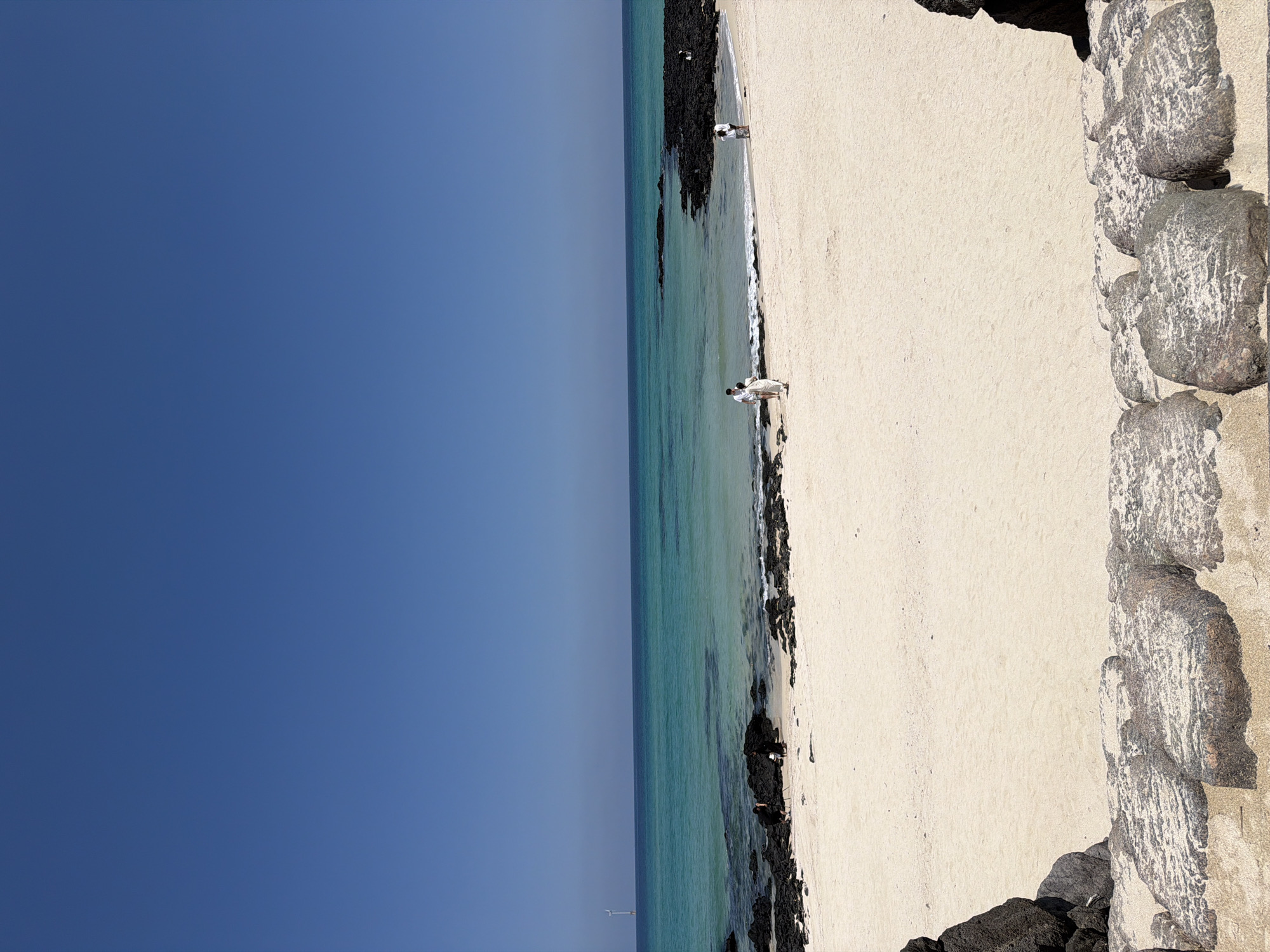 This image depicts a tranquil beach scene on Jeju Island, South Korea. The composition is divided diagonally, with a vast expanse of clear blue sky occupying the left half of the frame. The right half showcases a sandy beach meeting the turquoise waters of the ocean.
Several people are visible on the beach. In the foreground, two individuals in white attire are standing near the water's edge, appearing to engage in a conversation or activity. Further up the beach, another person in similar white clothing is also present. The presence of these figures suggests a leisurely day at the beach, possibly for recreation or a photoshoot given their matching outfits.
The beach itself is characterized by light-colored sand and a border of dark, rugged volcanic rocks, typical of Jeju's coastal landscape. The ocean exhibits a gradient of blues and greens, indicating clear and shallow waters. The sun appears to be high in the sky, casting shadows and suggesting it's daytime, likely late morning or early afternoon, with clear weather conditions. In the distance, a white structure resembling a wind turbine can be seen on the horizon, adding a modern element to the natural scenery. The overall mood is peaceful and serene, capturing the beauty of a sunny day at a Korean coastal destination. No visible text is present in the image.