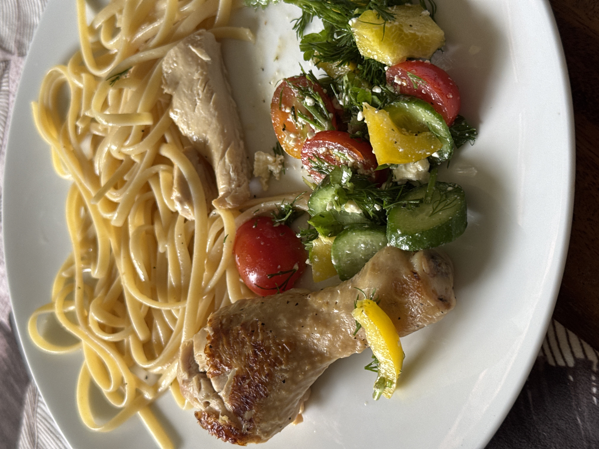 A close-up, overhead shot displays a plated meal consisting of pasta, chicken, and a fresh salad, served on a round white ceramic plate. The plate is positioned on a table with a light, striped fabric on the left side and a dark wooden surface on the right.
On the left side of the plate, a generous serving of light-colored pasta, resembling linguine or fettuccine, is coated in a glistening, light sauce, possibly creamy or oil-based, with hints of herbs. Beside the pasta, a piece of what appears to be cooked chicken breast, with a pale, soft texture, rests.
The right side of the plate features a colorful salad composed of halved red cherry tomatoes, sliced green cucumbers, and irregularly shaped pieces of yellow bell pepper. The salad is generously garnished with fresh green dill and sprinkled with crumbled white cheese, likely feta, and a light dressing.
Below the salad, a well-browned chicken drumstick is placed, showing some crispy skin and seasoning. A thin, bright yellow lemon wedge sits on the drumstick, suggesting a citrus flavoring.
The lighting is bright and natural, indicating daytime, and casts subtle shadows, highlighting the textures and freshness of the food. There is no visible text in the image. The scene depicts a prepared meal, possibly for lunch or dinner.