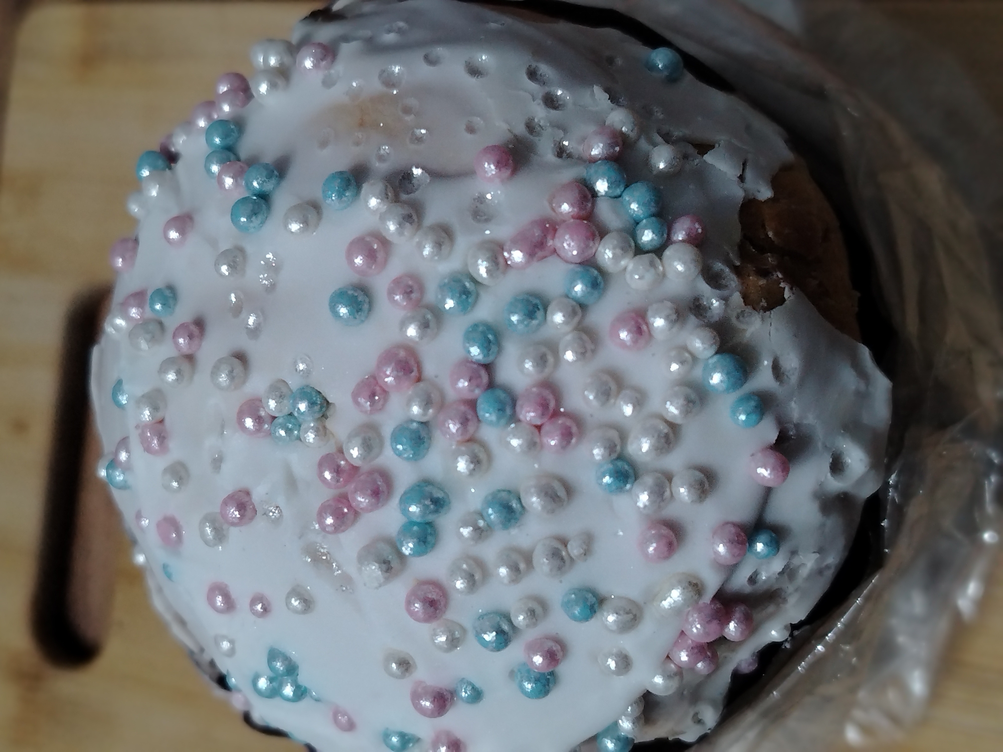 A close-up overhead shot depicts a Kulich, a traditional Russian Easter bread. The bread is topped with a thick layer of white icing and decorated with pink, white, and blue pearl sprinkles. The Kulich is partially enclosed in a clear plastic bag. The background is a light-colored wooden surface. There is no indication that this image was taken in Izhevsk, Russia. There are no people visible in the image.