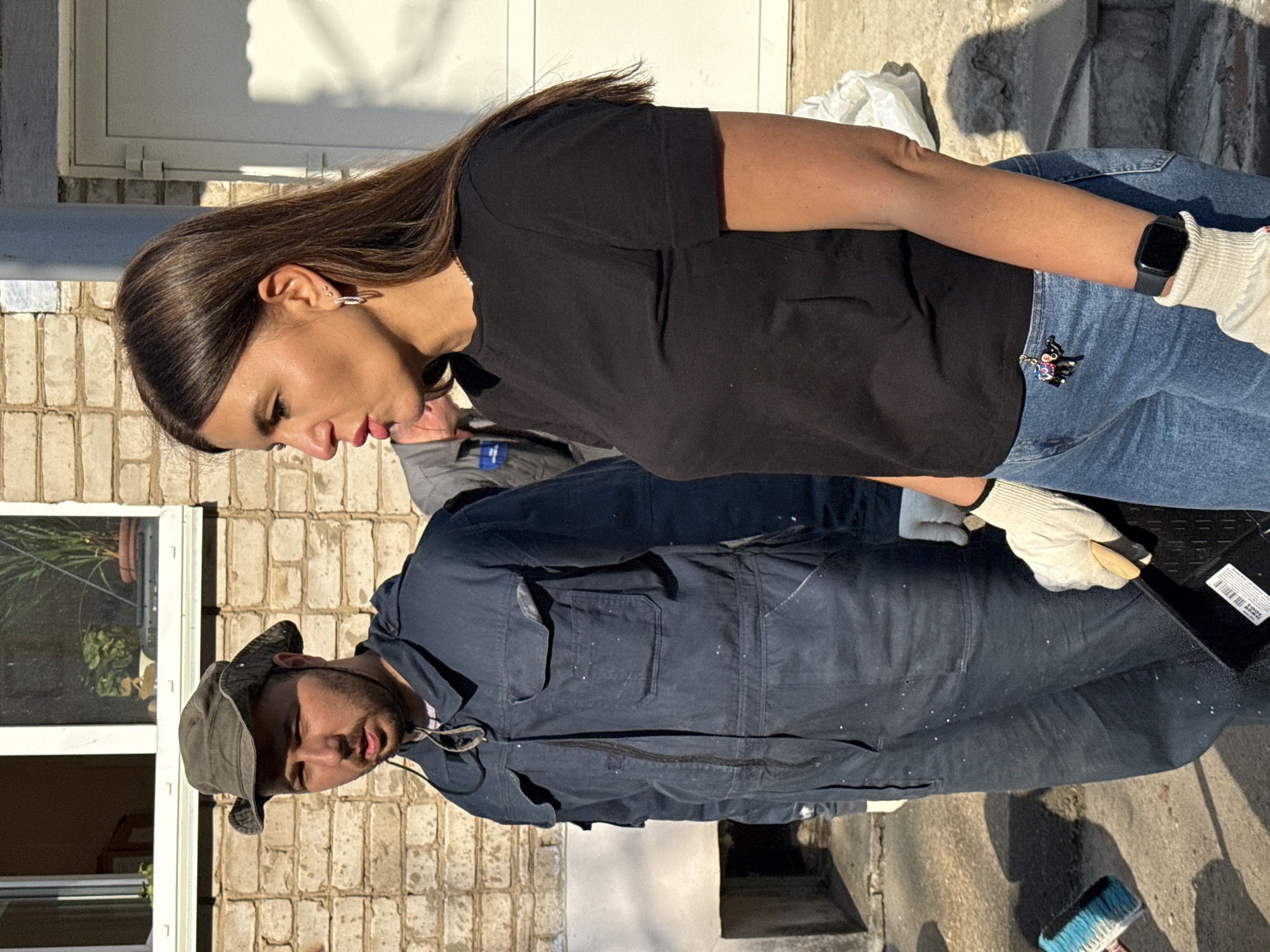 Outside, two individuals are engaged in cleaning. A woman with long brown hair, wearing a black t-shirt, blue jeans, and white gloves, is looking down. She has an Apple Watch on her left wrist and a small decorative pin on her jeans. A man wearing a dark blue work uniform and a bucket hat is also looking down, likely assisting in the cleaning task. A broom is visible near the bottom of the frame, suggesting sweeping is involved. The background shows a brick building with a window, indicating an urban setting. Given the prompt, the location is presumed to be Chaikovsky, Russia. The specific activities and relationship between the two individuals are not clear from the image alone, but they are collectively involved in some form of cleaning.
