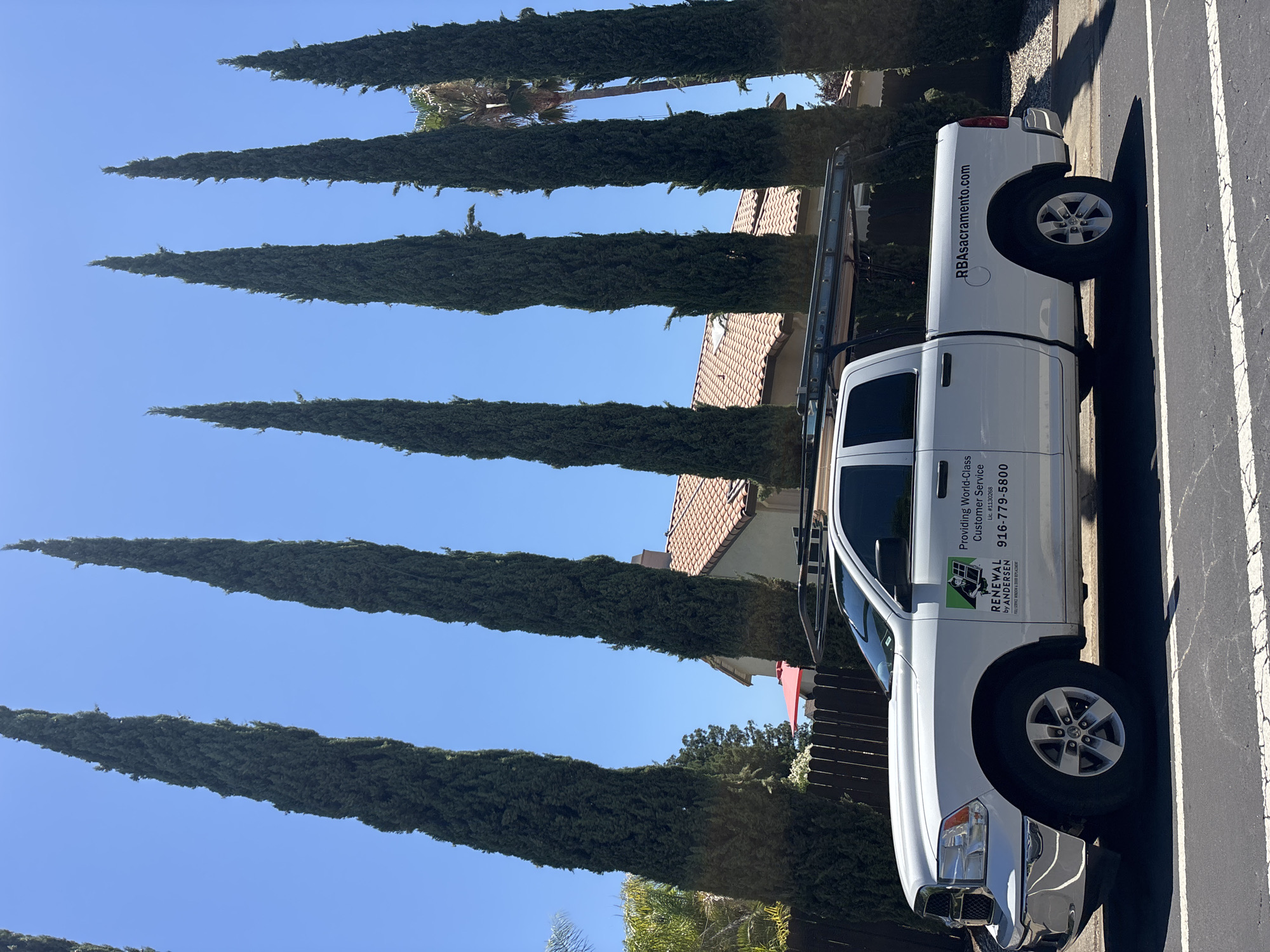 A white Dodge Ram pickup truck is parked on a residential street in Folsom, California. The truck has a ladder rack mounted on the roof and is branded with a logo and text that reads "RENEWAL by ANDERSEN" and "RBasacramento.com". The truck is parked in front of a house with a terracotta tile roof, bordered by a dark wooden fence and tall, narrow evergreen trees. The sky is clear and blue, and the sun is casting shadows.