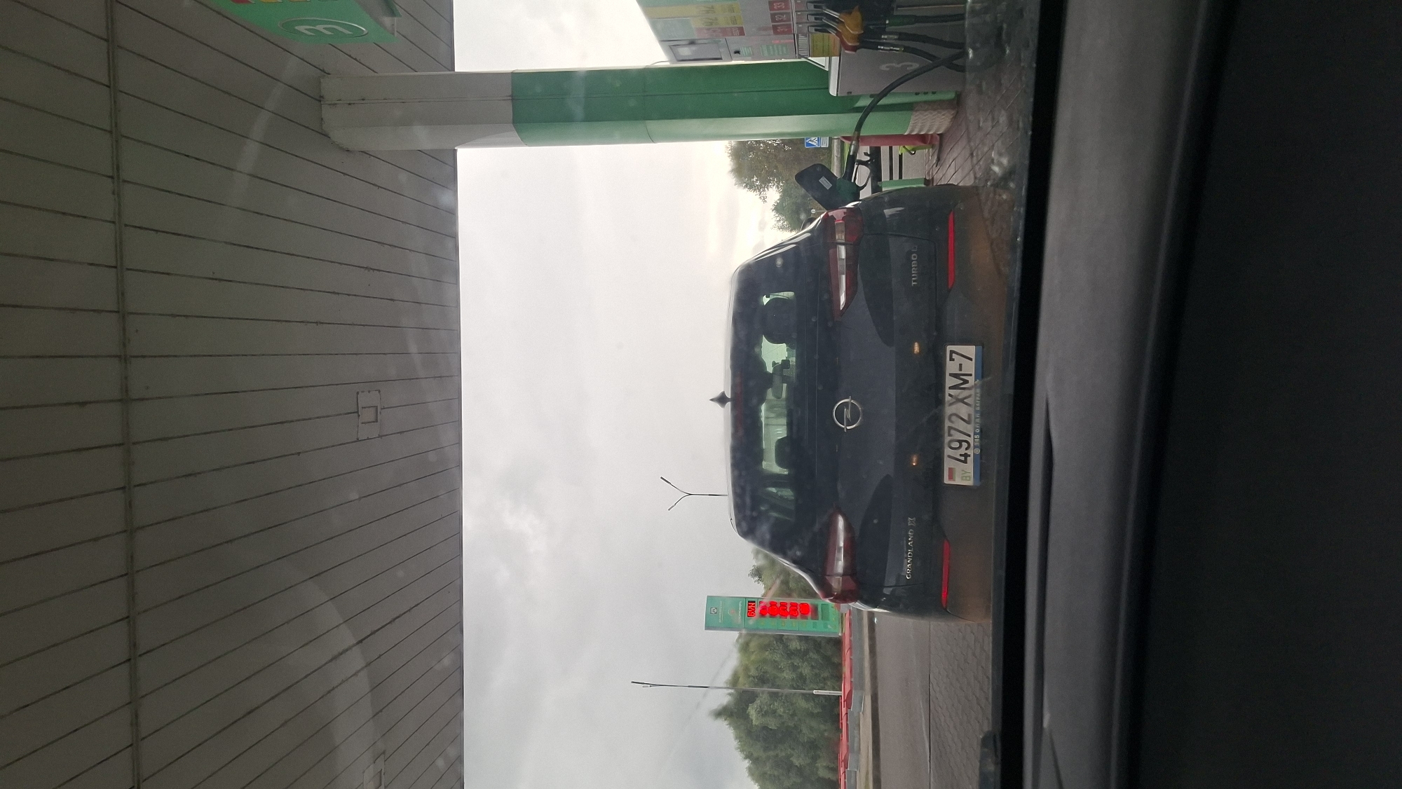 The image shows the rear of a dark colored Opel Grandland X car at a gas station in Myadzyel, Belarus. The car has a license plate that reads "4972 XM-7 BY". A gas pump hose is inserted into the car's fuel port. The gas station canopy is visible above the car. In the background, a digital sign displays fuel prices and trees are visible. The sky is overcast.