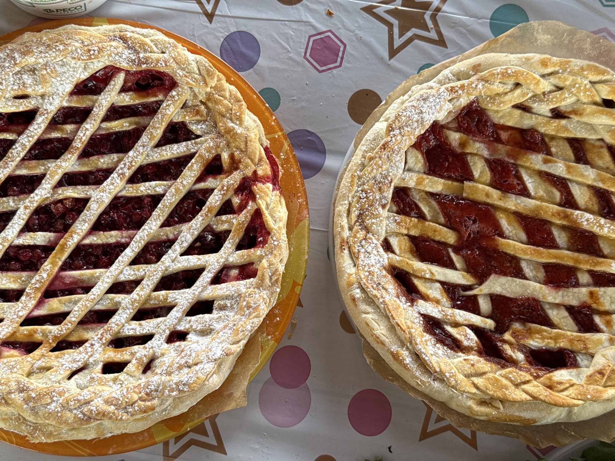 Two berry pies with lattice crusts dusted with powdered sugar sit side-by-side on a table covered with a patterned tablecloth featuring circles, hexagons, and stars. The pie on the left is on an orange plate. The location is not discernible from the image.