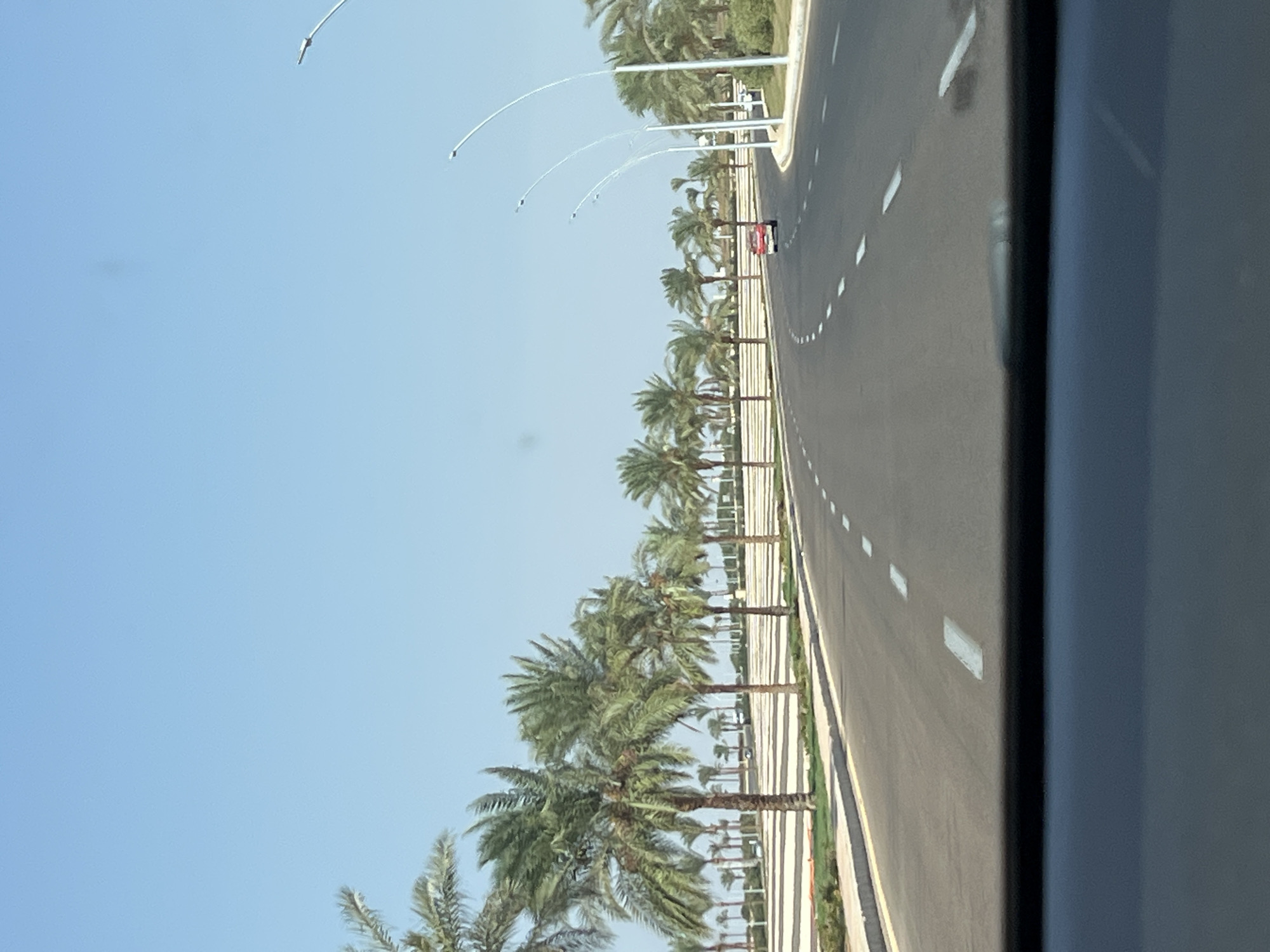 The image depicts a wide, paved road curving to the right, lined with numerous palm trees on the left and right sides. The road has white dashed lane markings and a yellow line along the right edge. A red car is visible driving on the road in the distance, on the right side of the lane. Modern, curved streetlights are also present along the right side of the road. The environment appears to be an urban or suburban area with well-maintained landscaping. The sky is clear and blue, suggesting a sunny day. This scene is likely located in Sharm El Sheikh, Egypt, a city known for its resort areas and palm-lined avenues.
