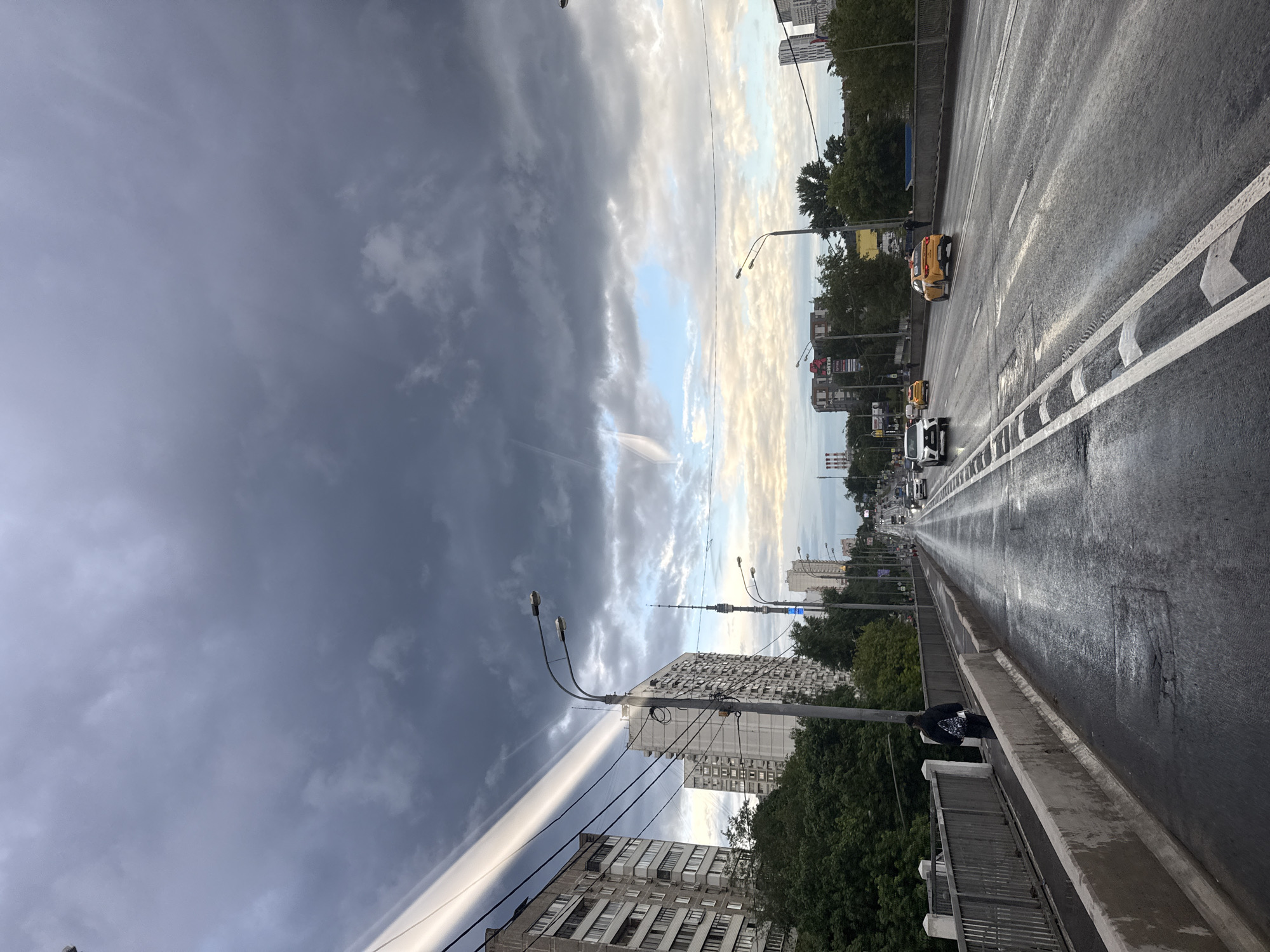 The image depicts a view of a city street in Moscow, Russia, under a dramatic, cloudy sky. The street is wet, suggesting recent rain, with visible reflections of the sky and surrounding buildings on its surface. Several cars, including yellow taxis, are driving on the multi-lane road. On the left side of the road, a pedestrian with a backpack walks along a sidewalk next to a metal railing. Buildings, some appearing to be apartment blocks, line the street, interspersed with trees. Streetlights are present along the road. A distinctive band of bright light, possibly sunlight breaking through the clouds, cuts diagonally across the sky on the left side of the image. The overall atmosphere is overcast and somewhat somber, characteristic of a cloudy day in an urban environment.
