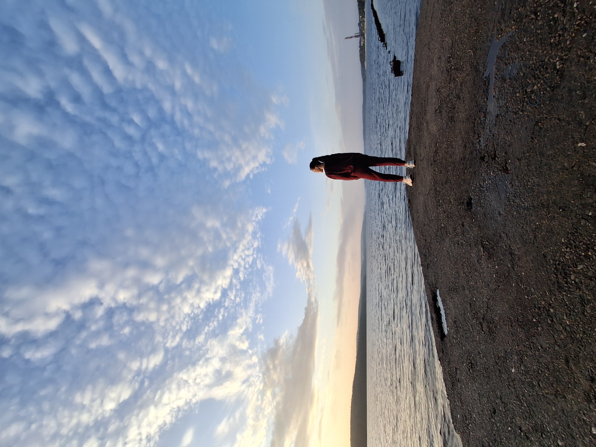 A person in a dark red tracksuit stands on a concrete pier overlooking a large body of water. The person is facing away from the camera, looking towards a distant horizon. The sky is a vibrant blue with scattered white clouds, and the sun appears to be setting or rising, casting a warm glow on the horizon. The water is calm, with gentle ripples. In the distance, a dark silhouette of hills or land is visible, and to the right, a long line of buoys or submerged objects extends into the water. A tall, industrial-looking structure is also visible on the far shore. The pier is rough and textured, with scattered pebbles and some puddles of water. The scene suggests a peaceful and serene moment by the water. This appears to be the Kama River in the city of Chaikovsky, Russia, specifically near the river dam or a similar waterfront area.