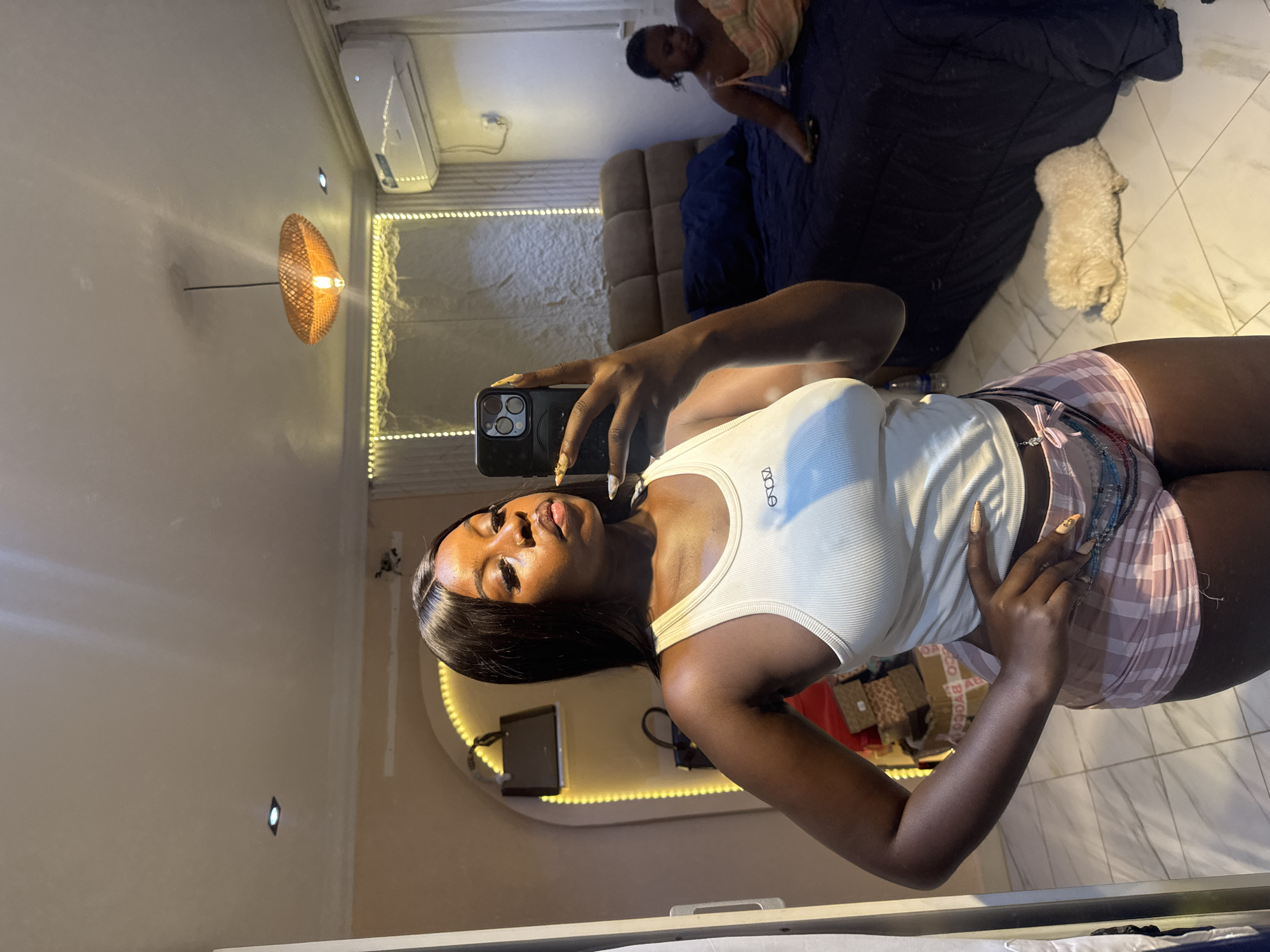 The media file captures a young woman taking a selfie in a well-lit bedroom. She is wearing a white ribbed tank top and pink plaid shorts. Her hair is long and black, and she has on full makeup, including dramatic eyeliner and lipstick. She is holding a black smartphone in her right hand.
In the background, reflected in a large mirror, another person is lying on a bed. There is a small, light-colored dog on the bed as well. The room has a modern aesthetic with white walls and a tiled floor. A woven pendant light fixture hangs on the left side of the frame, and a strip of warm LED lights runs along the wall. An air conditioning unit is visible on the upper wall. The overall mood appears relaxed and casual, likely during the evening or night given the interior lighting. The location is indicated as Abuja, Nigeria. No visible text is present on clothing or objects, other than a logo on the woman's tank top which is partially obscured.