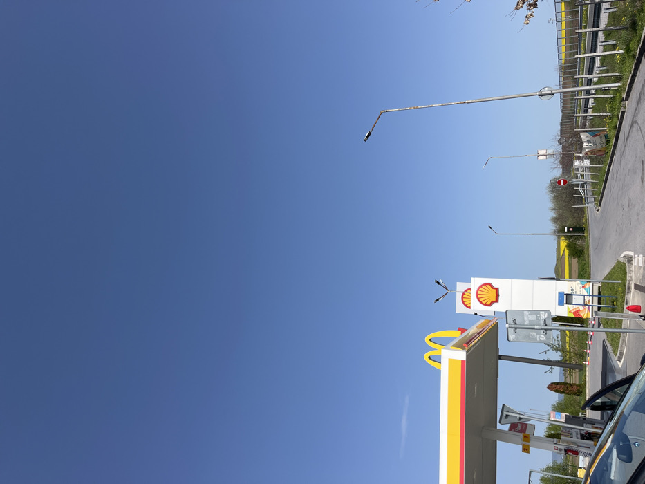 The image shows a Shell gas station in Hadjidimitrovo, Bulgaria, located at 42.532 latitude and 26.4 longitude. The gas station is on the side of a busy road, with a McDonald's logo visible on the roof of the canopy. There is a single white van parked in front of the gas pumps. There are several street lights on the road and a traffic sign displaying a red circle with a diagonal line through it. The sky is clear and blue with no clouds. The ground is paved asphalt with a small green grassy area surrounding the gas station. There is a green, grassy median strip separating the lanes of traffic.