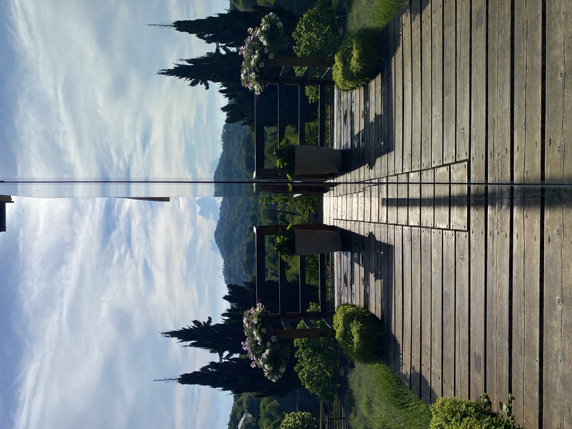 The image depicts a wooden deck leading towards a reflective glass partition, creating a mirrored effect of the surrounding landscape. The deck is constructed from weathered wooden planks, with visible nail heads and some water spots, suggesting it is outdoors. On either side of the deck, small, manicured green bushes are placed. Beyond the deck, a wooden railing encloses an area with two large potted plants and a potted tree adorned with white and pink flowers. In the background, several tall, dark cypress trees stand against a backdrop of rolling green hills and mountains under a partly cloudy sky. The scene is bathed in sunlight, casting shadows on the deck. The overall impression is one of a serene, well-maintained outdoor space in a natural, hilly environment. The image is likely taken in Verkhneye Uchdere, Russia, given the context provided.