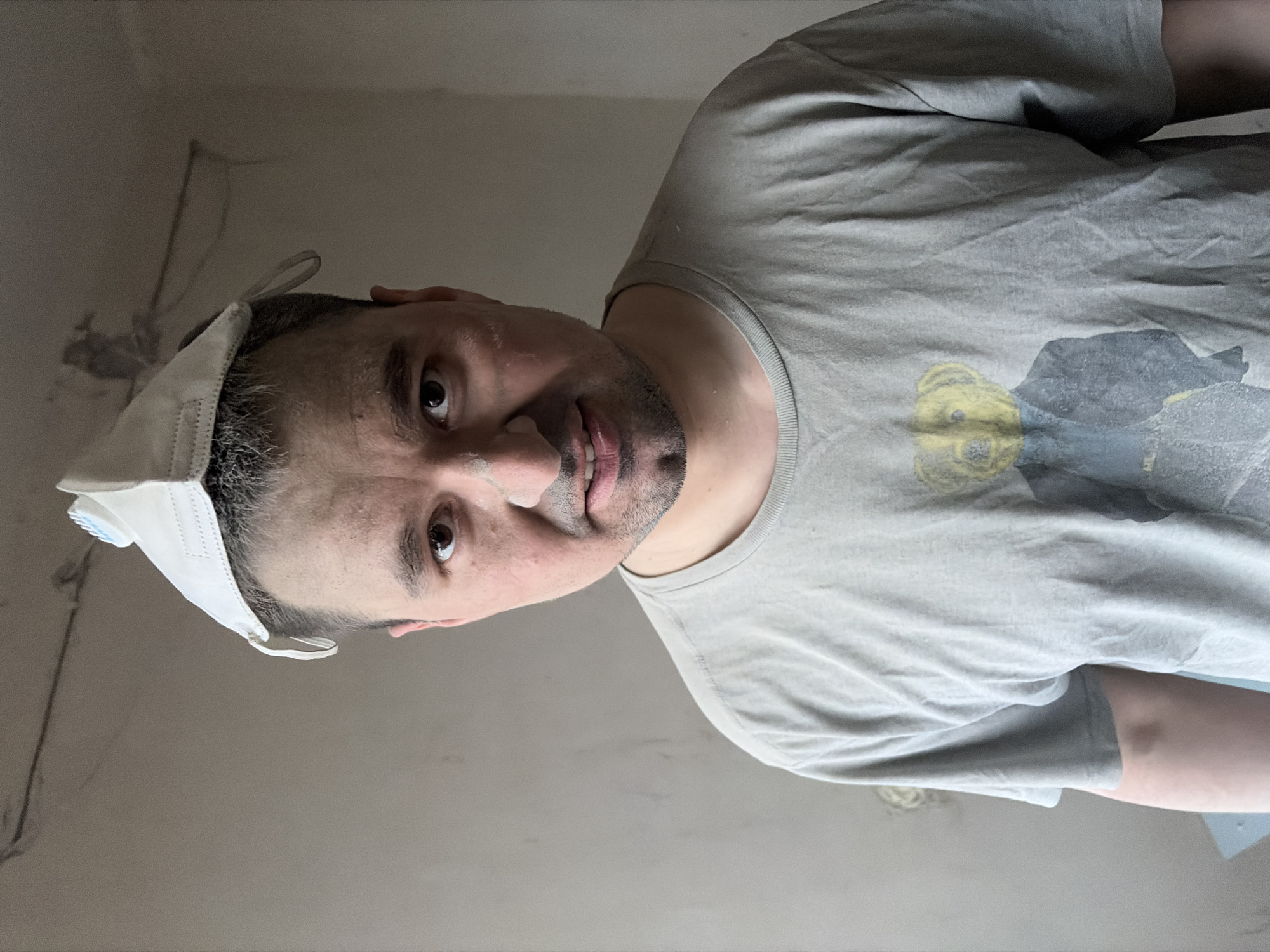 A middle-aged man with short dark hair and stubble is shown close-up, wearing a grey t-shirt with a faded bear graphic. He has a white face mask incorrectly positioned on the top of his head. The background is a pale grey wall, possibly indoors, with visible wiring. The city of Izhevsk, Russia, is not discernible from the image.