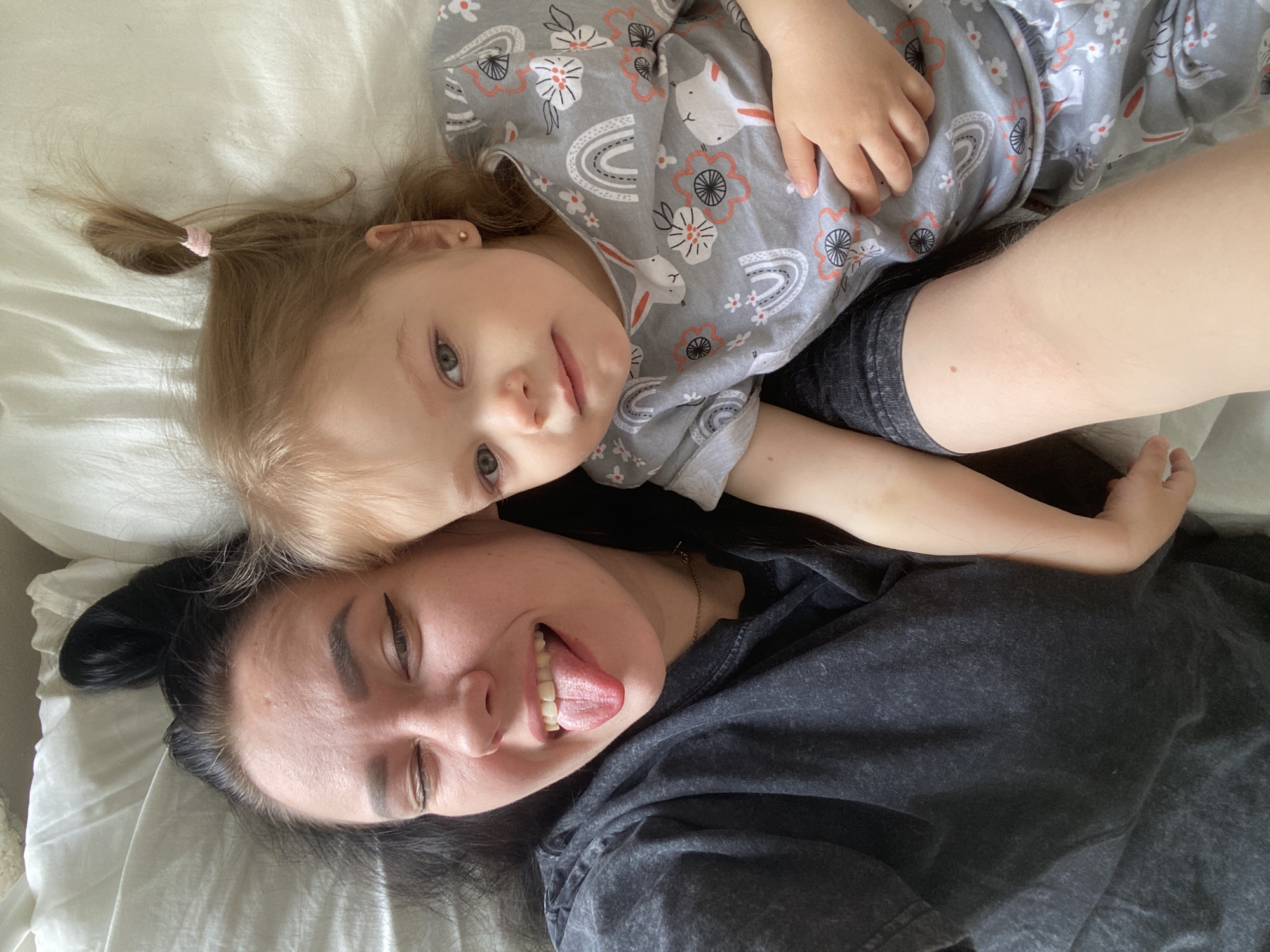 The image depicts a child and an adult lying on a white surface. The child, wearing a gray long-sleeved shirt with rabbit and flower patterns, is positioned on top of the adult, who is wearing a dark gray shirt. The child has blonde hair styled in a ponytail with a pink hair tie. The adult has dark hair pulled back in a bun. The adult's tongue is sticking out. The location is Unknown, Unknown.