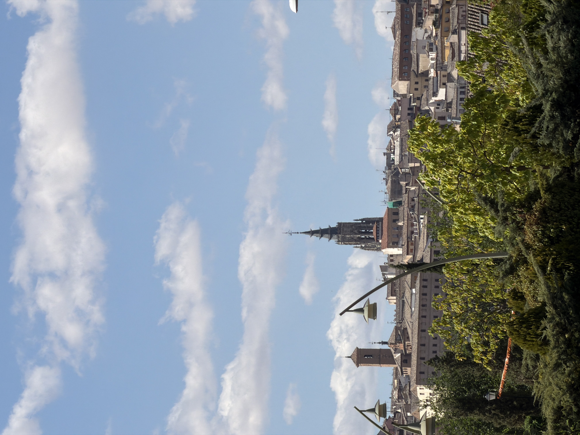 The image depicts a panoramic view of the city of Toledo, Spain, dominated by its historic architecture and lush greenery. In the foreground, a dense array of trees and shrubs creates a natural frame for the cityscape. The foliage is varied, with some trees bearing vibrant green leaves and others appearing more textured and dark. Interspersed within the greenery are two lampposts with distinct, bell-shaped fixtures.
The middle ground is occupied by the rooftops of Toledo, characterized by their terracotta tiles and a mix of architectural styles. Several buildings are visible, including what appears to be a church or cathedral tower with a pointed spire, and a separate square tower with arched windows. These structures suggest the rich historical and religious heritage of the city.
The background is defined by a clear blue sky dotted with fluffy white clouds, indicating fair weather. The overall scene is one of a well-preserved historic urban landscape set against a backdrop of natural beauty, likely captured from an elevated vantage point overlooking the city. The lighting suggests it is daytime, with the sun casting shadows and illuminating the buildings and foliage. No people are visible in the image.