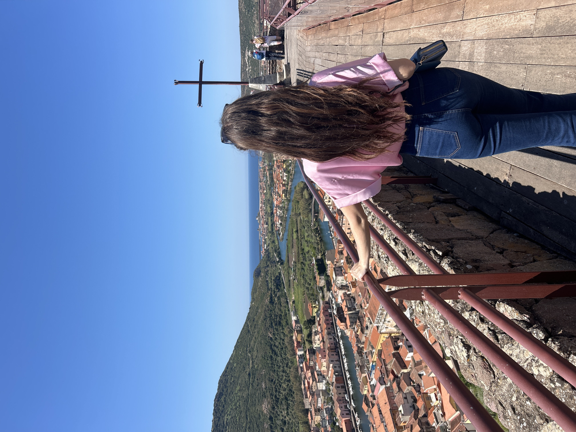 The image, oriented vertically, captures a person from behind, looking out over a panoramic landscape under a clear blue sky. The individual has long, wavy brown hair and is dressed in a light pink short-sleeved top and dark blue jeans. Sunglasses are visible on top of their head, and a small blue pouch or handbag is tucked into their waistband.
The person is positioned on a wooden platform, appearing to lean on dark red metal railings that separate them from the view below. In the immediate foreground beneath the railings, a stone wall is visible. Further back on the platform, other individuals can be seen in the distance, also observing the surroundings.
The expansive view showcases a picturesque town with numerous buildings, predominantly featuring warm-toned, likely terracotta, tiled roofs. The town is built along the winding course of a river or canal, where several boats are moored. Beyond the urban area, a large, lush green hillside slopes downwards, eventually giving way to the vast expanse of the blue sea meeting the horizon. A prominent feature in the sky is a simple, dark metal cross, situated against the clear azure.
The bright sunlight and distinct shadows suggest a sunny day, likely midday or early afternoon. The scene evokes a sense of peaceful contemplation and appreciation for the beautiful scenery. The location depicted is Bosa, Italy. No visible text is present in the image.