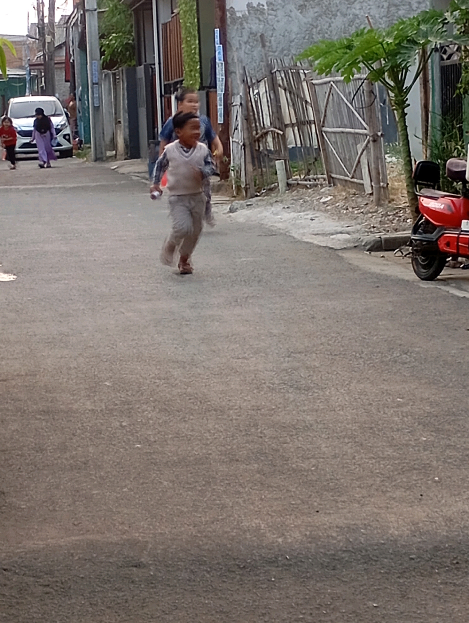 In Babelan, Indonesia, a young boy in a light-colored vest and gray pants runs down a paved street. He is accompanied by another child, who is out of focus behind him. In the background, a white car is parked on the side of the road. A red scooter is also visible, parked near a wooden fence. The scene appears to be a residential area with buildings and vegetation.