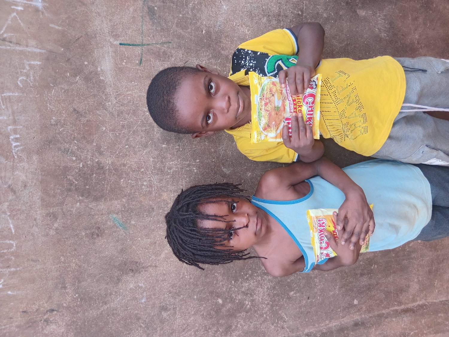 Two children are depicted lying on a concrete surface, likely outdoors, in Shagamu, Nigeria, during the daytime under bright natural light. The child positioned at the top of the frame has short dark hair and wears a yellow short-sleeved shirt with black and green patterns, along with grey athletic shorts. This child holds a large yellow packet of "Indomie Chicken Instant Noodles," clearly displaying the brand name and product type.
Below, the second child has dark dreadlocks and is dressed in a light blue sleeveless tank top with blue trim, paired with dark trousers. This child holds a smaller yellow packet, also identified as "Chikin Instant Noodles." Both children are looking directly at the camera with neutral expressions. The concrete ground beneath them is covered with various chalk markings, including numbers like '21', letter-like symbols, and a cross.