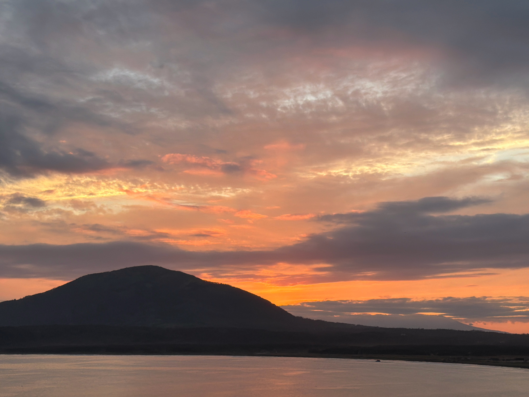 A silhouetted mountain rises from the dark landmass in the lower half of the image. The mountain's peak is rounded, and its slopes descend towards a dark, flat foreground that appears to be land. The foreground transitions into a body of water reflecting the colors of the sky. Above the land and water, the sky is filled with dramatic clouds. The clouds are a mix of dark grays, purples, and vibrant oranges and pinks, indicating a sunset or sunrise. The lighting suggests either early morning or late evening. The scene is devoid of people or animals. This is a scenic view of a natural landscape, possibly the coast of Yuzhno-Kurilsk, Russia, given the reference to the city, featuring a prominent mountain and a body of water under a colorful sky.