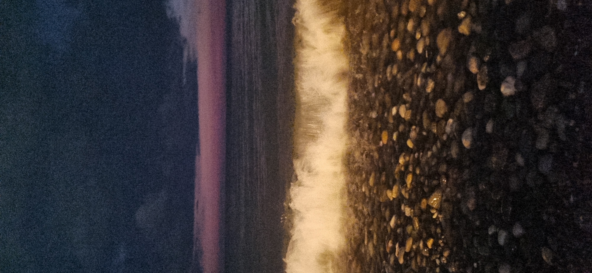 This is a photograph of a stony beach at dusk, likely in Batumi, Georgia. The sky is a deep, dark blue, transitioning to a soft pink band just above the horizon, indicating either sunrise or sunset. Below the sky is the dark expanse of the sea, with the faintest suggestion of waves. In the foreground, a foamy wave is breaking onto the shore, illuminated by an unseen light source. The beach itself is covered in dark, wet pebbles and stones of varying sizes. The overall mood is tranquil and slightly dramatic, with the low light and crashing wave.