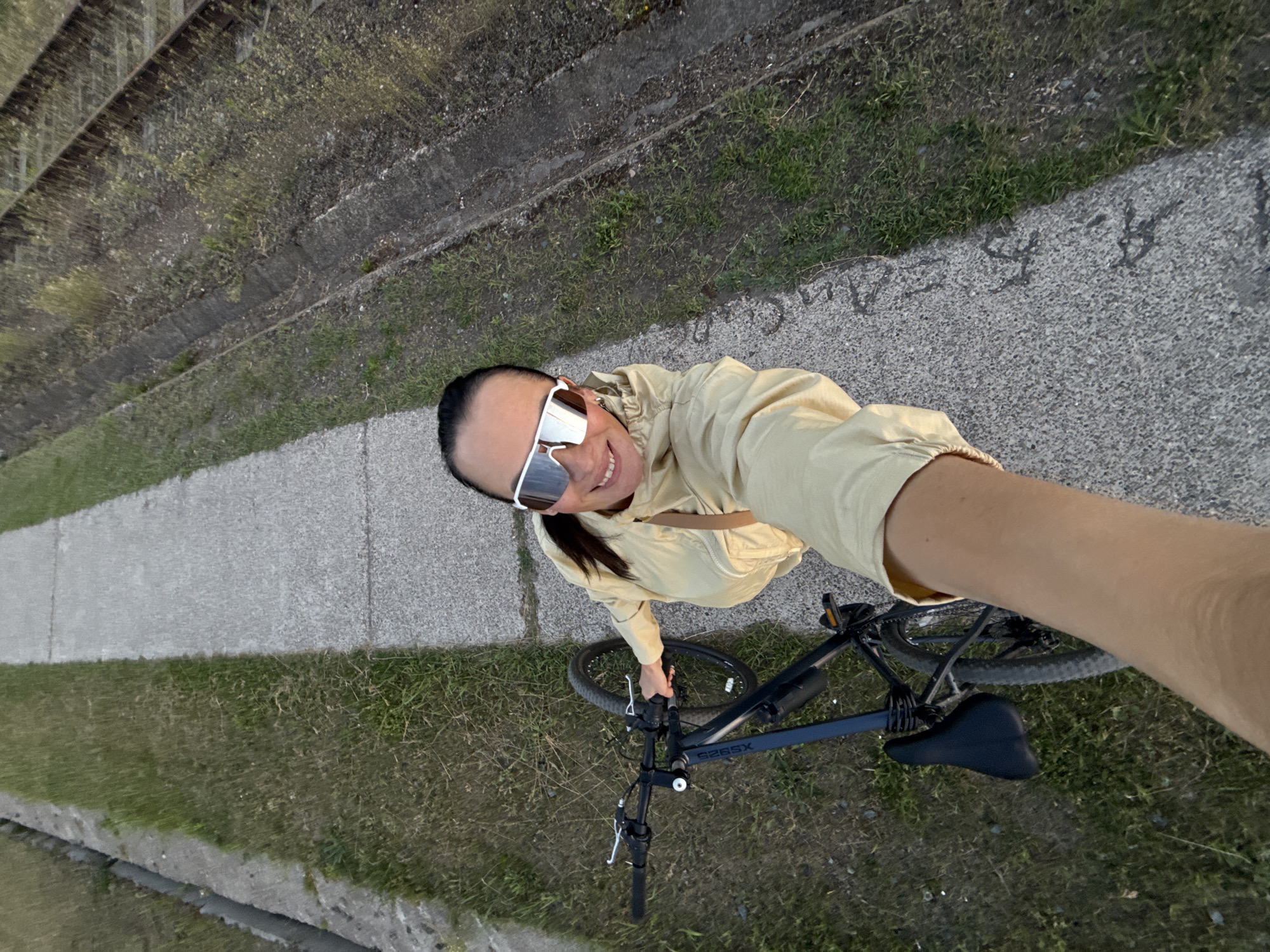 An overhead, wide-angle selfie shows a woman wearing a light yellow jacket and white mirrored sunglasses, smiling and holding a black bicycle by the handlebars. She is standing on a paved walkway next to a grassy area and what appear to be train tracks. The walkway is cracked and shows some discoloration. The grassy area is dry and sparse in some places. The train tracks are set in concrete and are overgrown with weeds. The scene appears to be an outdoor industrial or utility area. The woman's right arm and hand are visible at the bottom of the frame as she takes the photo. The background extends to show a long, straight walkway receding into the distance, with the grassy area and train tracks flanking it.