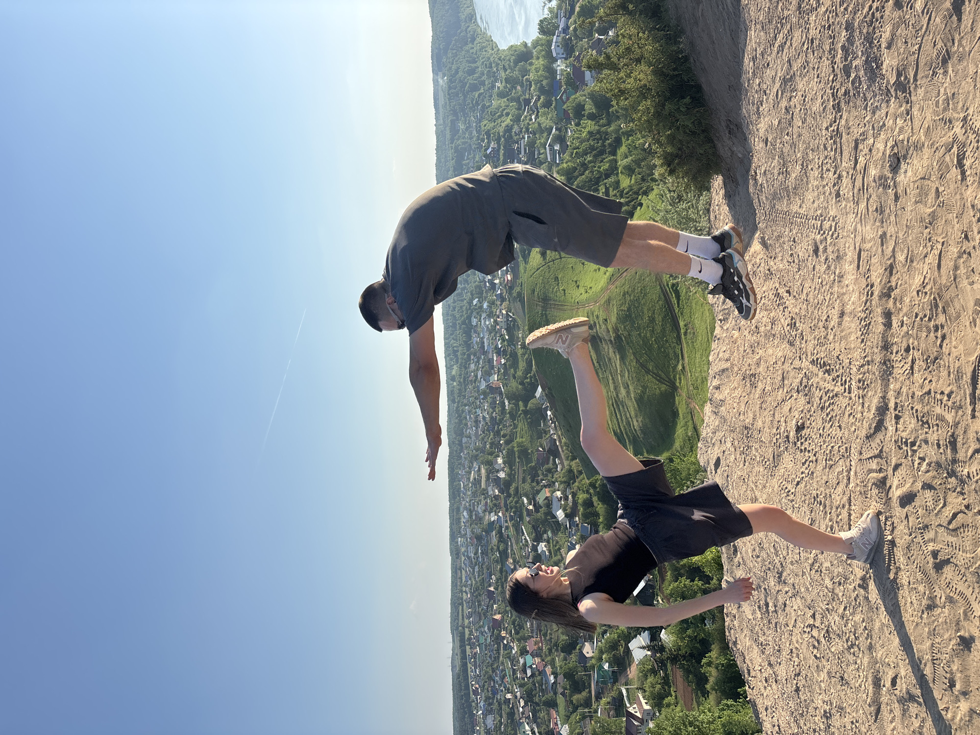 The image shows a man and a woman appearing to leap off the edge of a sandy cliff overlooking the city of Ольховка, Russia. The man, closer to the top of the frame, is positioned with his body bent forward as if diving, wearing a grey shirt and shorts with white socks and dark sneakers. The woman, positioned below him, is in a similar pose with one leg extended upward, wearing a dark tank top and shorts with light-colored sneakers. The background reveals a town with houses and greenery extending to the river, indicating the image was taken on a cliffside. The sky is a clear blue, and the ground near the edge of the cliff is sandy with numerous footprints.