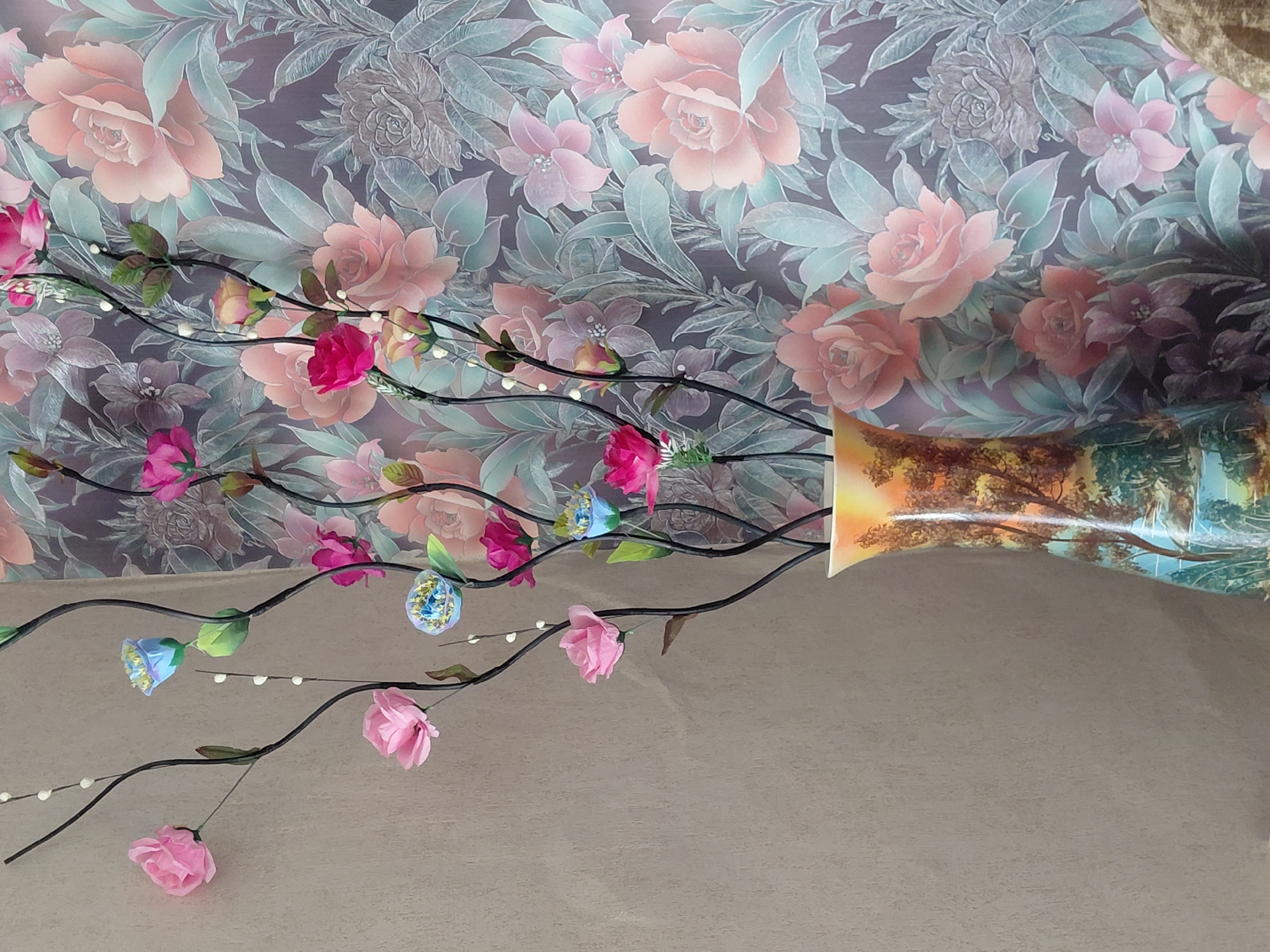 The image shows an artificial floral arrangement in a patterned vase, positioned against a floral wallpaper. The floral arrangement consists of dark, slender branches adorned with pink and blue artificial flowers, interspersed with small white beads. The vase is cylindrical with a flared opening and features a landscape painting with trees and a body of water. The wallpaper behind the arrangement displays a dense pattern of large, stylized flowers and leaves in shades of pink, purple, and green. The scene appears to be indoors, likely in a residential setting. There are no people visible in the image. The location is not specified as being in Novosyolovo, Russia.