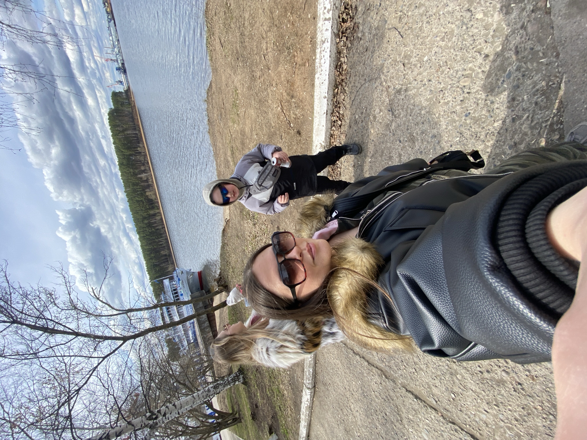 On a sunny day in Chaikovsky, Russia, two individuals are present near the Kama River. One person, wearing sunglasses, a black leather jacket with a fur collar, and long brown hair, is taking a selfie. Another person, wearing a gray hooded sweatshirt and sunglasses, is standing near the water holding a silver object, possibly a can. The background shows the Kama River, a line of trees, and cruise ships docked along the shore. The foreground includes a concrete path and a few birch trees without leaves. The sky is partly cloudy.