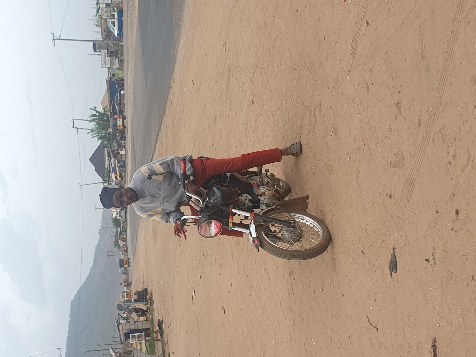 The image captures a daytime scene on a dusty, unpaved road, likely in a semi-urban or rural setting. The central subject is a young man, seated on a red motorcycle, looking directly at the camera with a neutral to slight smile. He is dressed in a light-colored, possibly grey and white, long-sleeved jacket, red trousers, and dark shoes, with a dark cap on his head. His right foot is placed on the ground, indicating he is either stopped or about to move.
The motorcycle is predominantly red and black, featuring chrome handlebars, a front headlight, and spoked wheels. The ground is reddish-brown earth, showing tire tracks and small scattered debris.
In the background, on the left side of the road, there are various modest buildings and what appear to be small shops or stalls. Several vehicles, including cars and "keke napep" (motorized tricycles), are visible along the roadside. Utility poles and power lines crisscross the upper part of the frame. A prominent, green-covered mountain range stretches across the distant background under an overcast sky. The diffused lighting suggests a cloudy day.
No discernible text is visible on any signs, vehicles, or objects within the image. The environment and vehicles are consistent with everyday scenes in Lokoja, Nigeria.