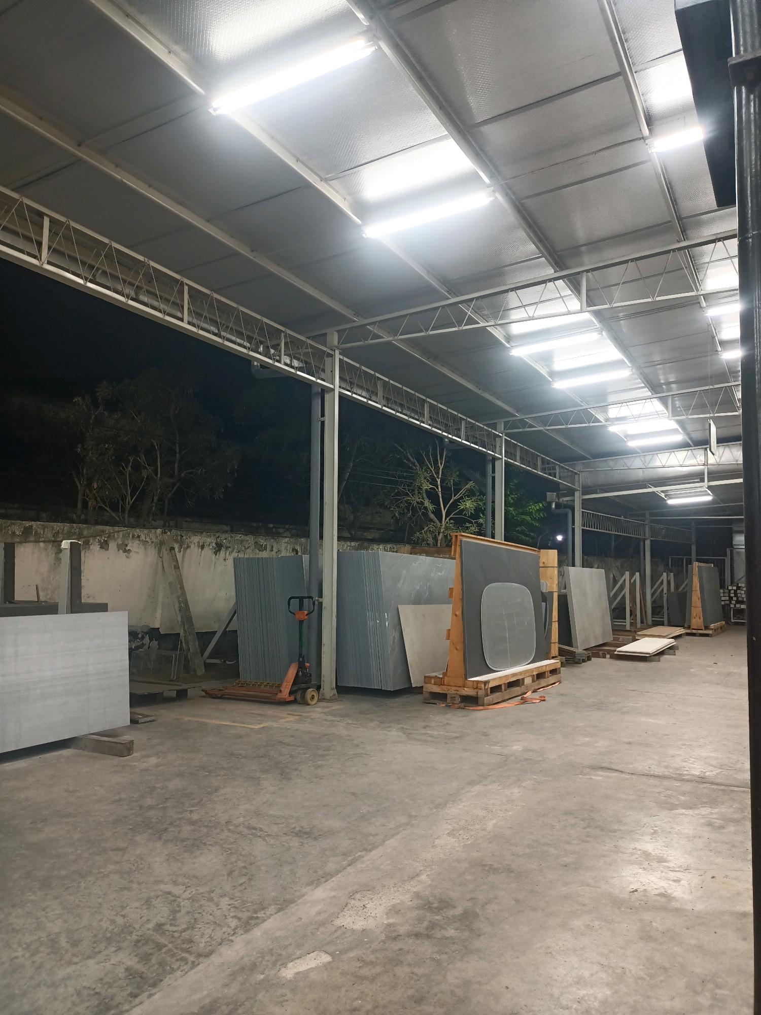 The image depicts an indoor warehouse or storage facility, likely at night given the external darkness and internal artificial lighting. The structure appears to be a covered area with a metal roof supported by beams and trusses. Fluorescent lights are mounted on the ceiling, illuminating the space.
The floor is a concrete surface, showing some signs of wear and tear with stains and cracks. The warehouse contains stacks of large, flat materials, possibly stone slabs or panels, arranged vertically and horizontally on pallets and stands. Some of these materials are gray, while others appear to be lighter in color.
In the foreground, to the left, is a large, light-colored slab resting on a low concrete support. Further into the scene, a bright orange manual pallet jack with black wheels is positioned near a stack of gray slabs.
In the middle ground and background, more stacks of slabs are visible, some on wooden pallets and others in wooden frames. One prominent display features a large, dark gray slab with an oval cutout, leaning against a wooden frame. Beyond the immediate storage area, the darkness outside suggests trees and a wall.
There are no people visible in the image. The overall impression is one of a well-lit, functional storage space for bulk materials. The location is identified as the city of Ngargosari, Indonesia.