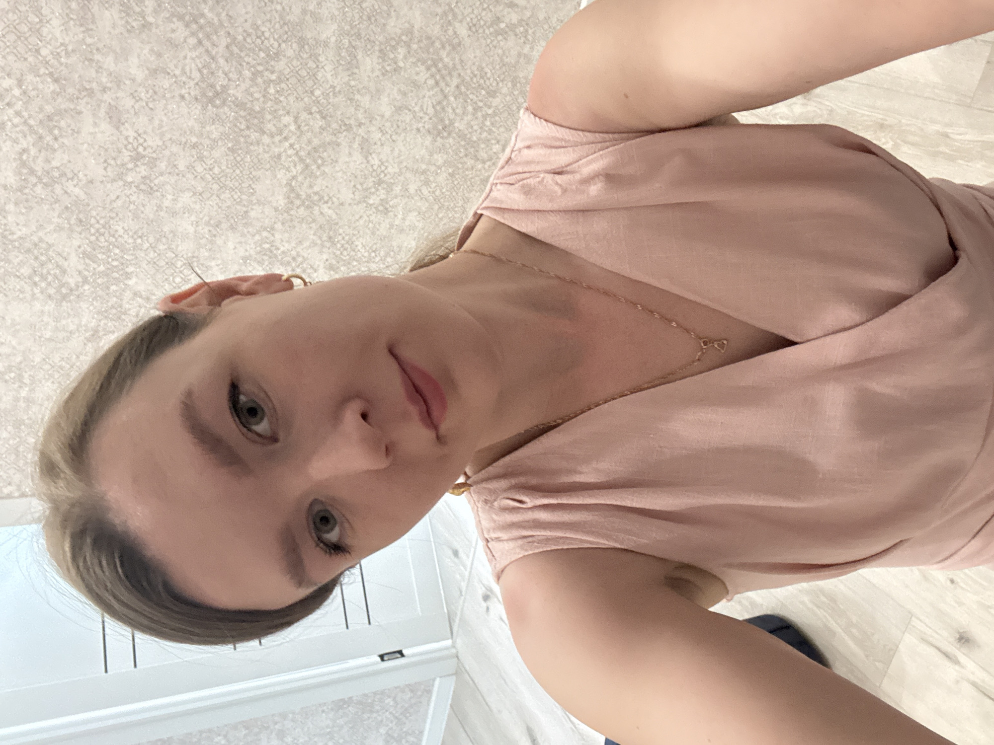A woman is seen in a selfie-style photo, her face oriented slightly downwards towards the camera. She has fair skin, green eyes, and blonde hair pulled back. She is wearing a light pink, wrap-style dress and a delicate gold necklace. The background includes a textured wall and a partially visible white door with vertical panels. There is no indication of Izhevsk, Russia, or any specific city location in the image.
