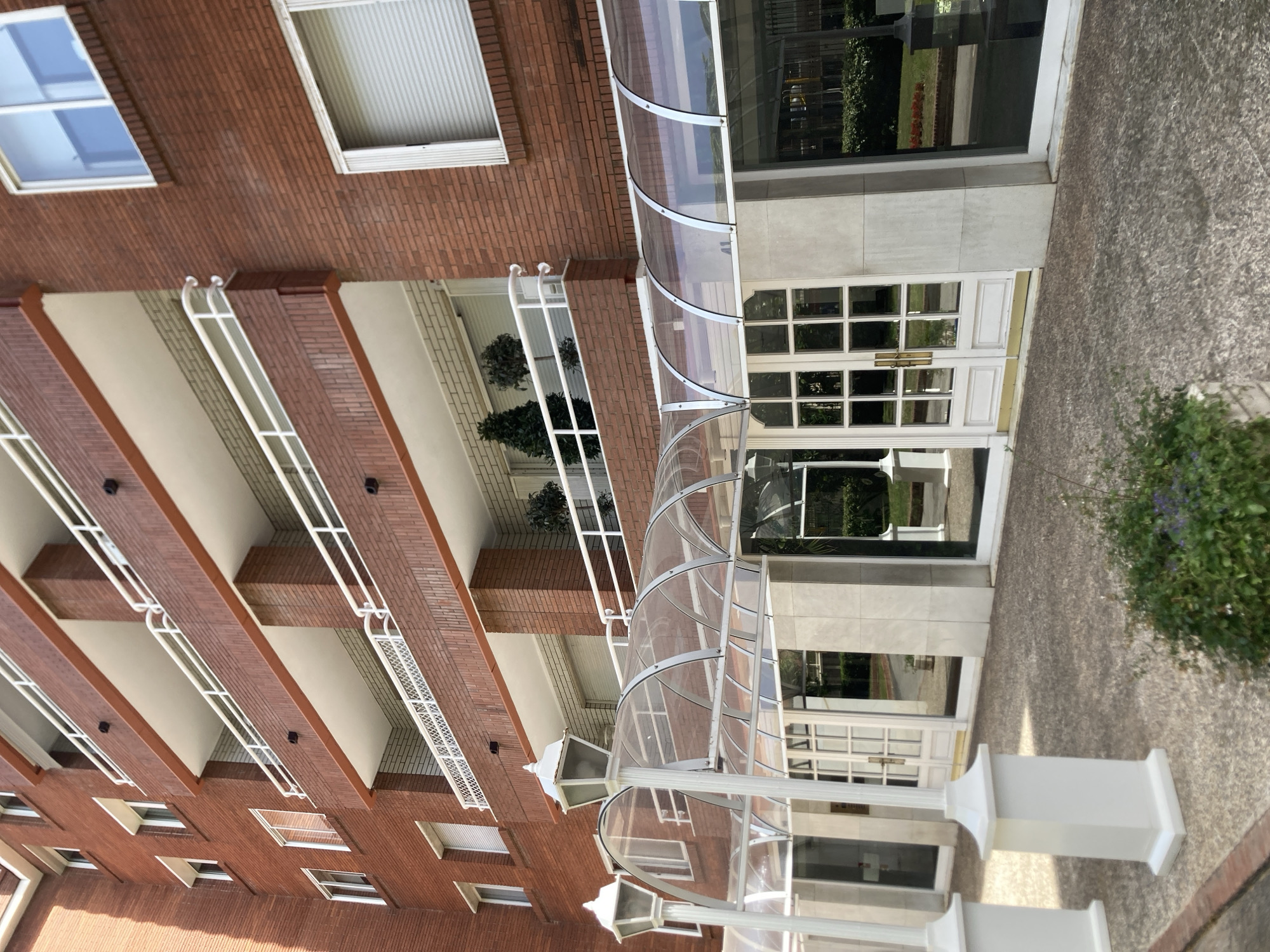 The image shows the facade of a red brick apartment building. The building has multiple floors with balconies that have white railings. The ground floor has a covered entrance with a clear plastic awning supported by white posts. There are white double doors with glass panels, and large windows. The pavement in front of the building is made of speckled concrete. A small patch of green bushes with purple flowers is visible in the foreground. The image does not contain any people or provide specific information about its location within Barcelona, Spain.