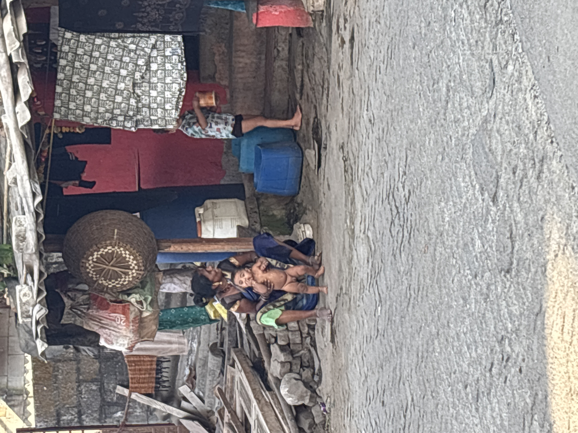 A woman is holding a baby next to a pile of discarded bricks and wood on a concrete surface. Next to them are personal belongings: cloth, bags, a basket, etc. A man lies on top of blue containers. Over him hang blankets and clothing. The location is in Ambernath, India.