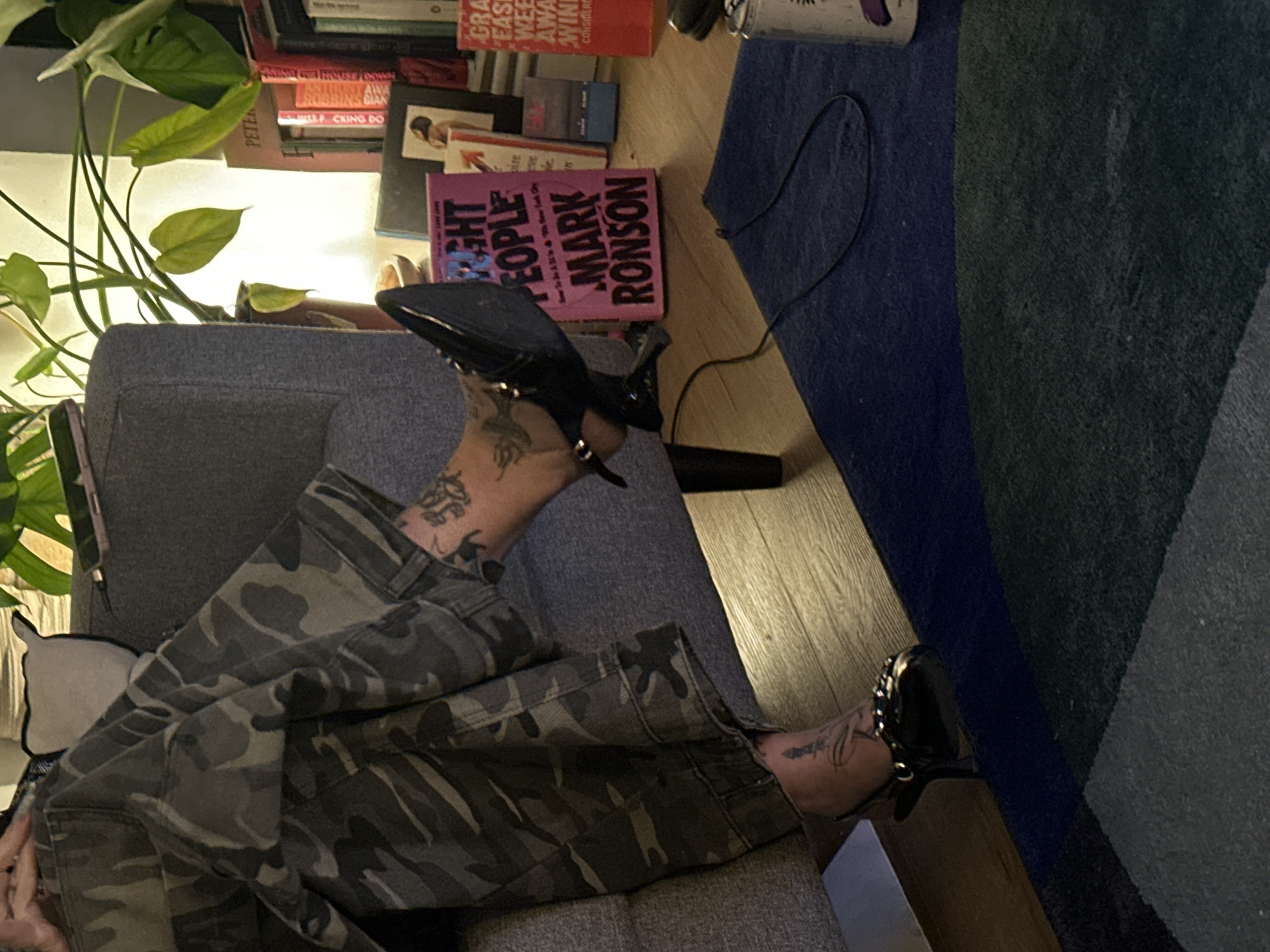 The image depicts a person sitting in a gray armchair. The person is wearing camouflage pants and black high-heeled shoes. The person has tattoos on both ankles. Next to the chair is a small bookshelf with several books, including "Right People" by Mark Ronson. A blue, gray, and green rug is on the wooden floor. A green potted plant is visible next to the bookshelf.