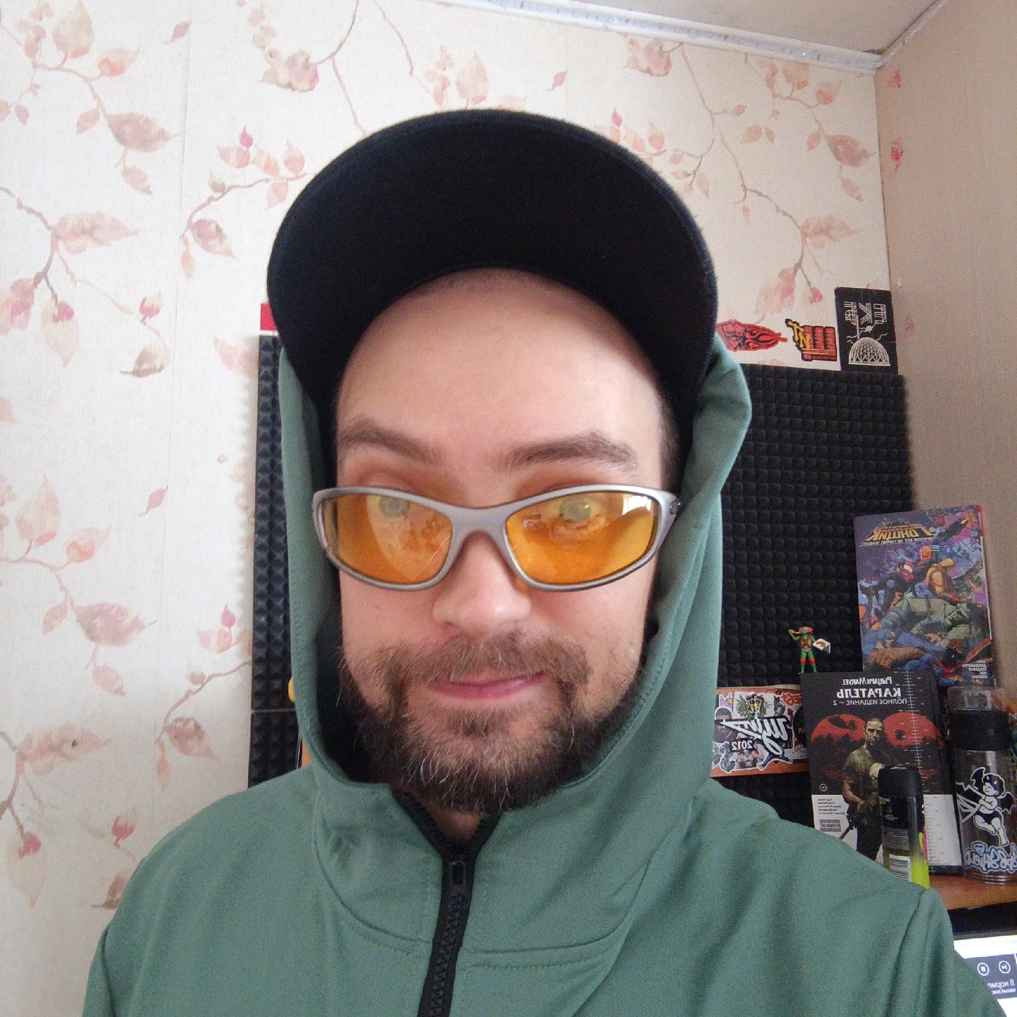 The image depicts a close-up, selfie-style shot of a person wearing a dark baseball cap and a green hooded jacket. They are also wearing silver-framed sunglasses with orange lenses. The person has a beard and mustache. The background features floral wallpaper and a wall with several stickers and a black acoustic foam panel. To the right, there is a stack of items including a book, a small figurine, and a metal container with stickers. The scene appears to be indoors. There is no visible indication or information in the image to suggest the location is Yekaterinburg, Russia.