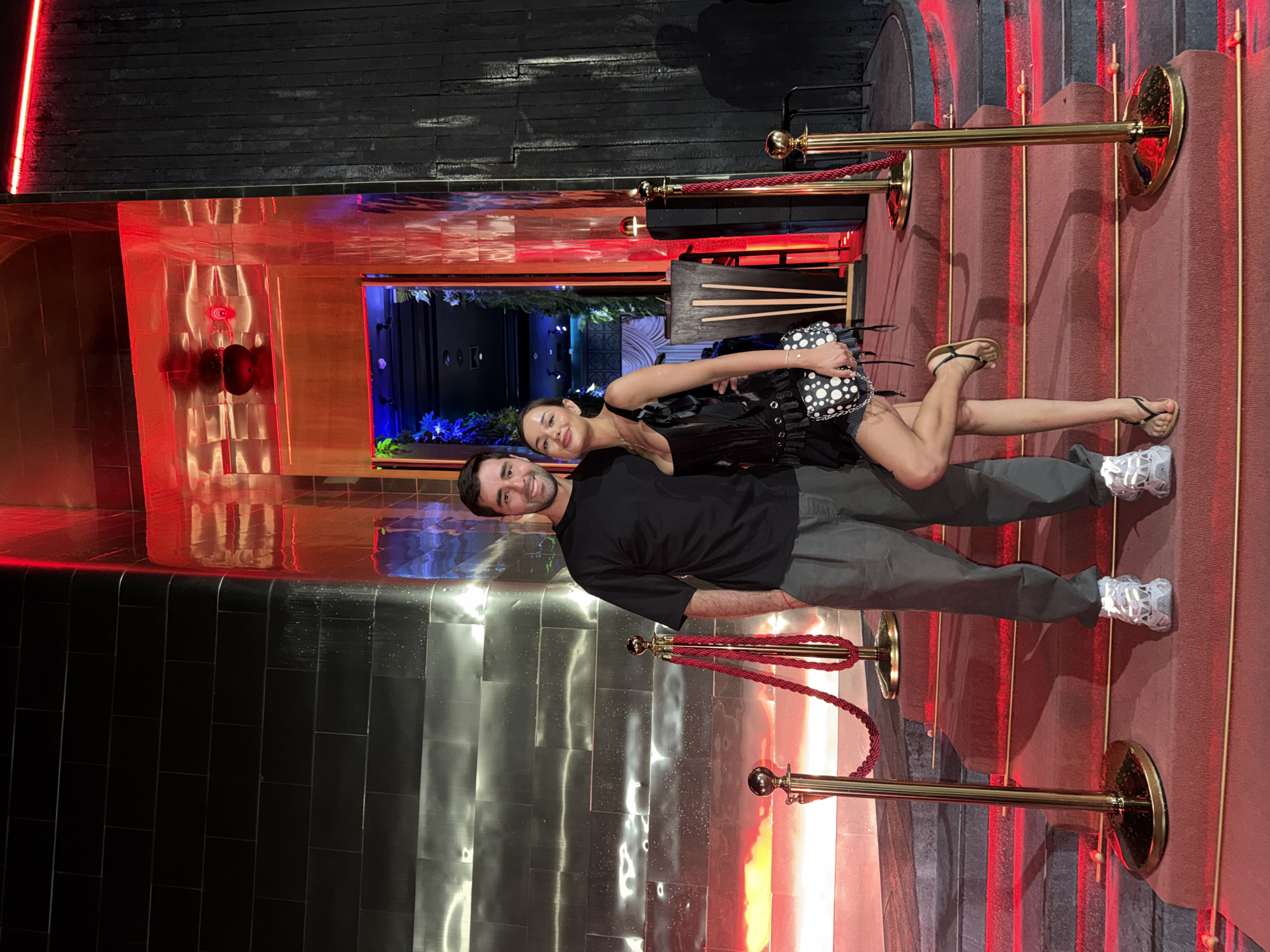 A man and a woman pose on a red carpet in front of a club or bar. The man is wearing a black T-shirt and gray pants with white sneakers. The woman is wearing a black corset and denim shorts with gold flip-flops, carrying a black and white polka dot purse. They are standing behind a velvet rope line. The building has a dark black tiled facade, red lighting, and a mirrored wall with red accents. The scene is illuminated with a combination of red and blue lighting. The location is in Choeng Thale, Thailand.