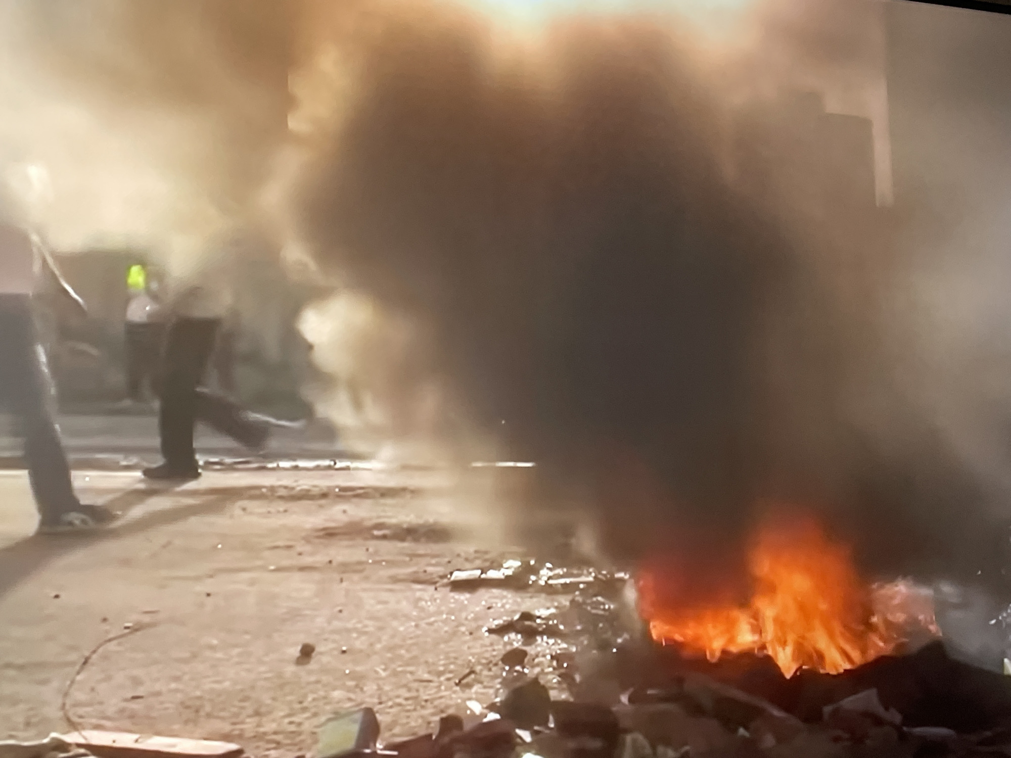 The image depicts a street scene in an unidentified city. A fire is burning on the right side of the street, producing thick smoke. Two individuals are walking on the left side of the street, away from the fire. Debris is scattered around the area of the fire. A building is faintly visible in the background through the smoke.