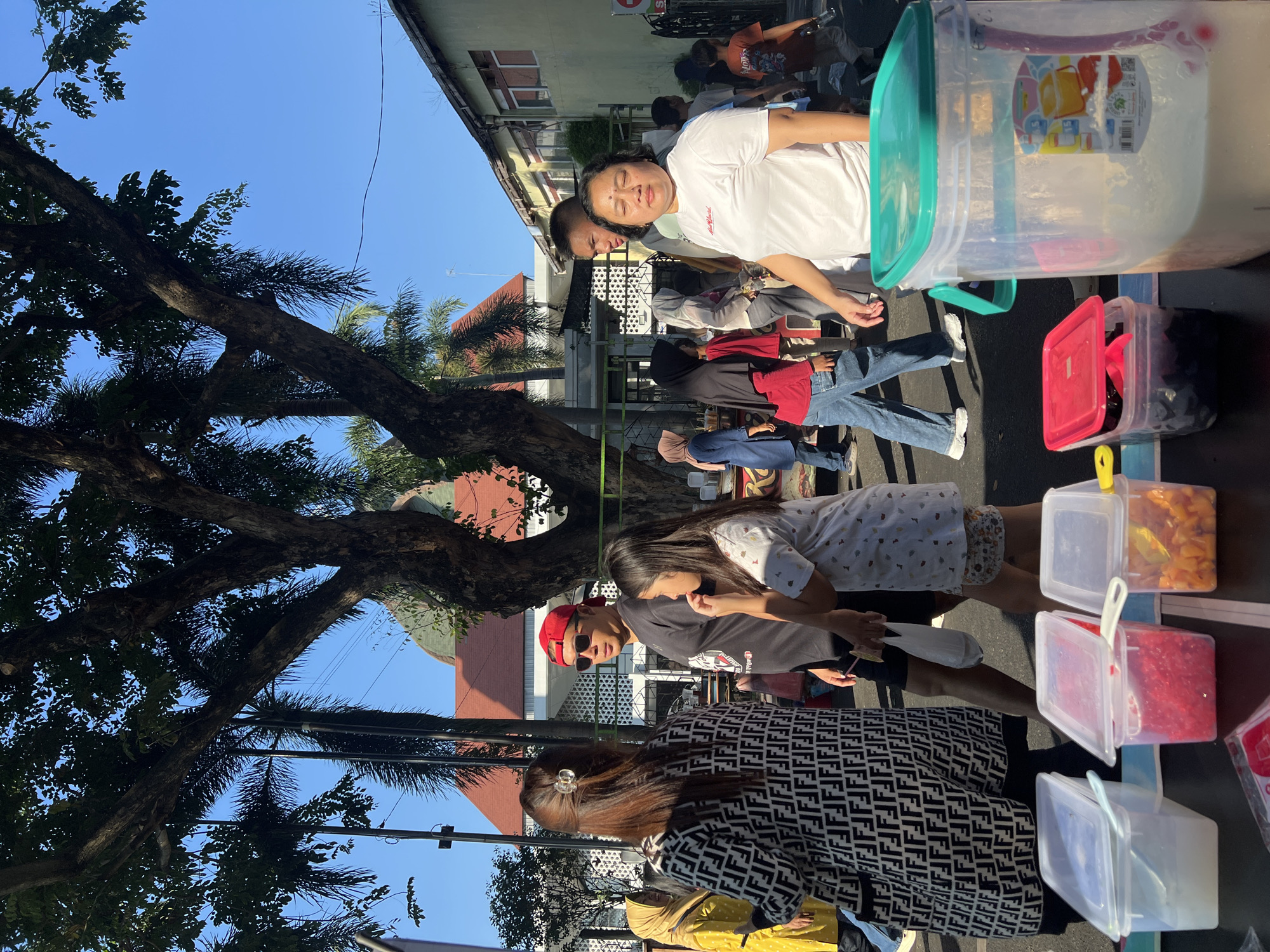 This image depicts a street scene in Kecamatan Pati, Indonesia, on a clear day. In the foreground, several transparent plastic containers hold colorful ingredients, likely for food or drinks. To the left, a person in a black and white patterned garment is visible from the waist up. In the center, a man wearing a red baseball cap and sunglasses is talking to a young girl. Further to the right, a woman in a white t-shirt stands looking forward. Behind them, a crowd of people are walking and interacting. Tall trees with lush green leaves provide shade, and buildings with traditional architecture, including a domed roof, are visible in the background. The overall atmosphere suggests a bustling outdoor market or event.