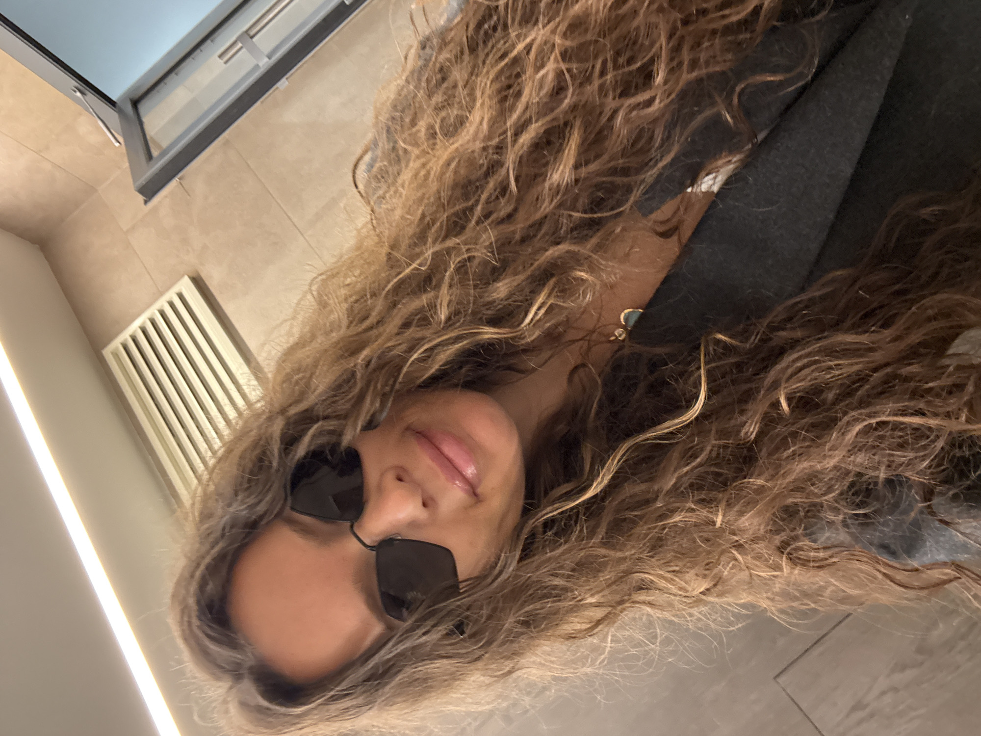 A young woman with long, curly brown hair and dark sunglasses is taking a selfie. She is wearing a dark blazer over a light-colored top and a necklace with a blue pendant. The background appears to be an indoor space with tiled walls, a ventilation grate, and a glass door. The lighting suggests it may be daytime. The woman is looking at the camera and smiling. Her lips are glossy. The overall impression is casual and relaxed. The location is specified as Khimki, Russia.
