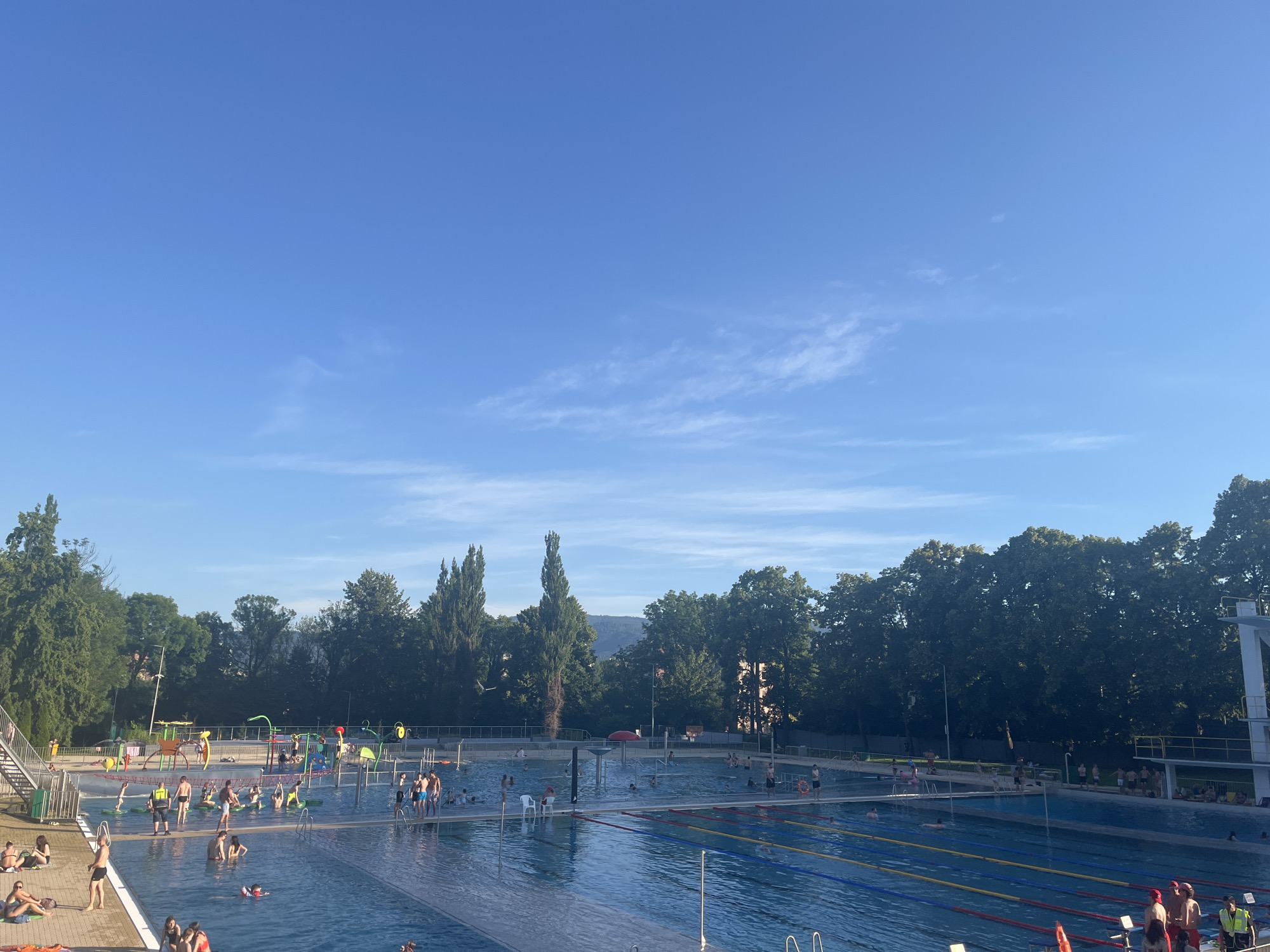The image depicts a sunny day at an outdoor swimming pool complex, likely located in a city park or recreational area given the abundant trees bordering the complex and the distant view of buildings and hills. The sky is a clear, bright blue with a few wispy white clouds.
The main focus is the large swimming pool, which is divided into sections. One section, on the right, features multiple swimming lanes marked with red, yellow, and blue lane lines, suggesting it's a competition or lap swimming area. This section also has a diving platform visible on the far right. In the foreground and midground, people are engaged in various water activities. Some are swimming, others are wading, and a few are lounging on the pool deck. There are also several children visible in a shallower area, which appears to be a splash pad with colorful playground equipment, including slides and water features.
Around the pool, there are people sunbathing on towels and lounging chairs on the paved deck. Trees line the perimeter of the pool complex, providing shade in some areas. The overall atmosphere is one of leisure and recreation, with a noticeable number of people enjoying the warm weather and the facilities. The scene is captured from an elevated perspective, looking down and across the pool area.
