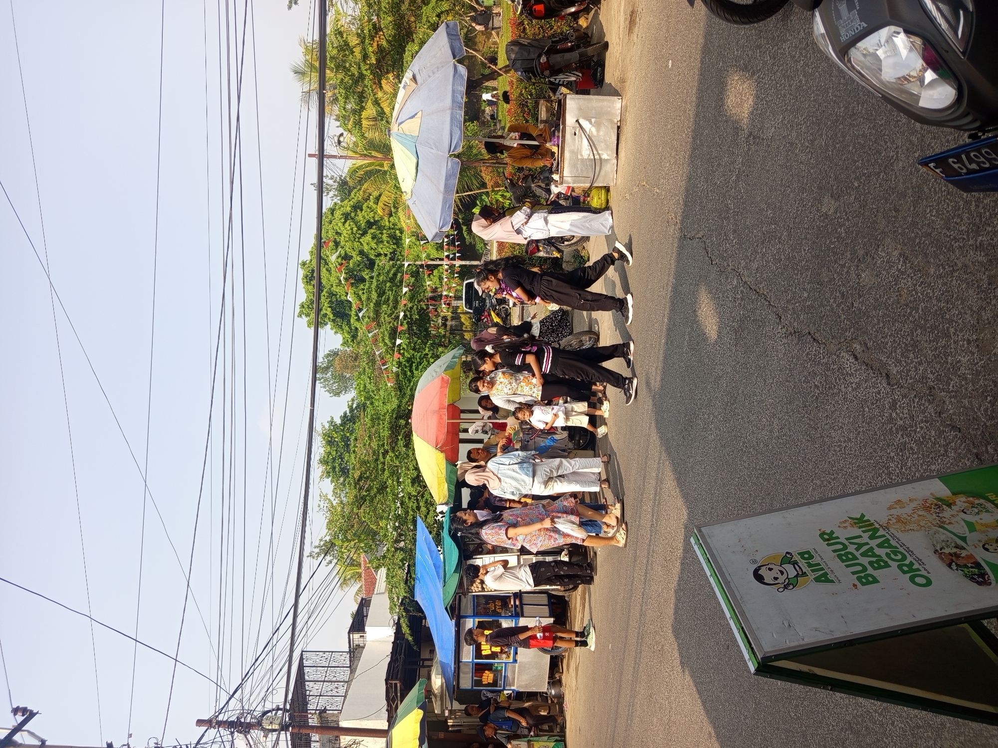 The image depicts a bustling street scene in Ciomas, Indonesia, likely during daytime given the strong sunlight and shadows. Several food stalls with colorful umbrellas are set up along the roadside. People of various ages are gathered around the stalls, some queuing, some socializing, and others appearing to purchase food.
In the foreground, a green and white A-frame sign displays the brand "Aira" and promotes "Bubur Bayi Organik" (Organic Baby Porridge), featuring images of the product. The street is paved with asphalt and shows some cracking.
Further down the street, several food carts are visible. One cart, under a blue awning, has signage in Indonesian. Another stall under a large blue, yellow, and green umbrella features a person cooking at a grill. Motorcycles are parked in the background, suggesting local transportation.
The environment includes lush green trees and vegetation lining the background, with a bright sky overhead. Overhead power lines crisscross the sky, a common feature in urban and semi-urban Indonesian landscapes. The overall atmosphere is that of a lively local market or street food gathering.