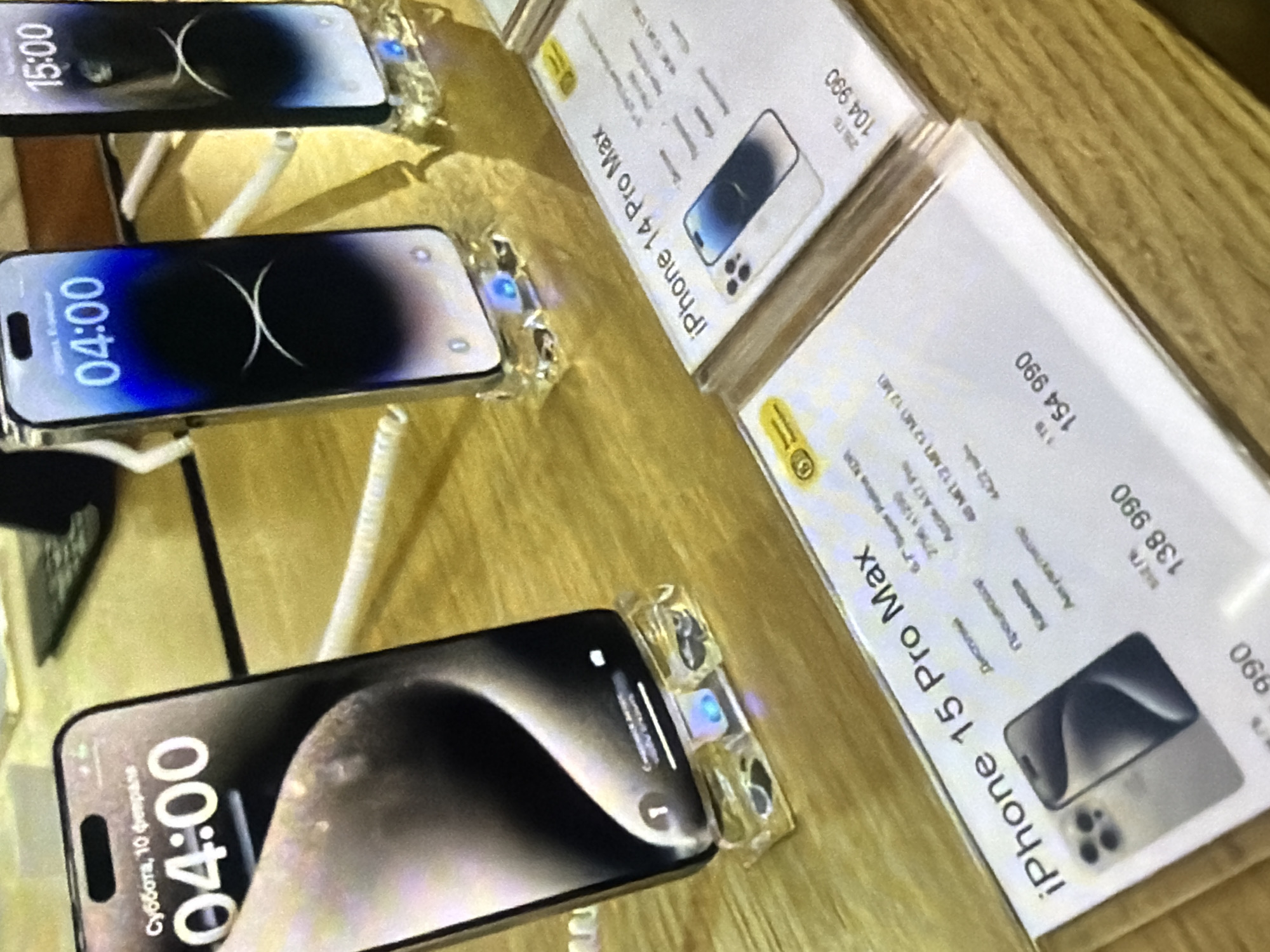 The image shows a close-up view of several iPhones displayed on a wooden surface. The iPhones are upright and positioned on clear plastic stands, with a white charging cable visible on the left. Three iPhones are visible in focus: one on the far left displaying "04:00" and "Суббота, 10 февраля" (Saturday, February 10th) on its screen. To its right, another iPhone also displays "04:00" on its screen, with a blue glow emanating from its base. A third iPhone on the far right displays "15:00".
In the foreground, there are two price tags or informational cards. The one on the left, labeled "iPhone 15 Pro Max", shows an image of the phone and details such as "6.7\" Super Retina XDR", "Apple A17 Pro", "48 Mpix 12 Mpix 12 Mpix", and "4422 mAh". It also lists prices for different storage capacities, with "138 990" clearly visible for an unspecified storage size. The card to the right of this one appears to be for an "iPhone 14 Pro Max" and shows a price of "104 990" for "256 GB". Another section of a card further to the right indicates a price of "154 990" for "1 TB".
The lighting in the scene is bright, likely from overhead store lighting, casting some shadows. The background is blurred, but appears to be a retail store environment with other electronic devices possibly present. The image's focus is on the iPhones and their accompanying product information. There are no people visible in the image. The location of Bukuru, Nigeria is not discernible from the visual information present in the image itself.