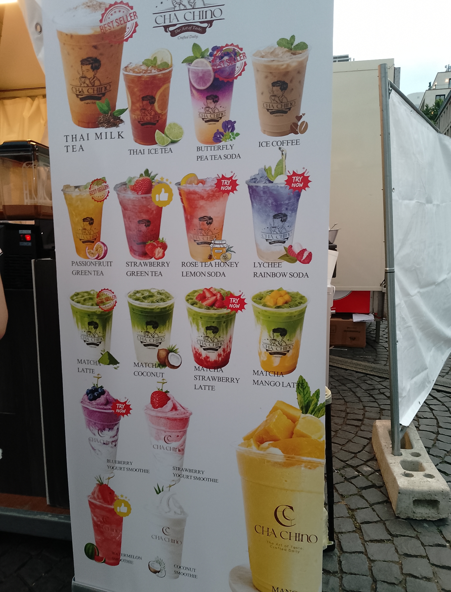 The image is a close-up, low-angle shot of a vertical menu board for "CHA CHINO The Art of Taste Crafted Daily". The menu displays various drinks with images and text. The top row features "THAI MILK TEA", "THAI ICE TEA", "BUTTERFLY PEA TEA SODA", and "ICE COFFEE". The second row showcases "PASSIONFRUIT GREEN TEA", "STRAWBERRY GREEN TEA", "ROSE TEA HONEY LEMON SODA", and "LYCHEE RAINBOW SODA". The third row includes "MATCHA LATTE", "MATCHA COCONUT", and "MATCHA STRAWBERRY LATTE". The bottom row features "BLUEBERRY YOGURT SMOOTHIE", "STRAWBERRY YOGURT SMOOTHIE", a "WATERMELON SMOOTHIE", and a "COCONUT SMOOTHIE". To the right of the menu board, a white tarp is visible, supported by a metal pole and a concrete block. The ground is paved with cobblestones. The lighting suggests it is daytime.